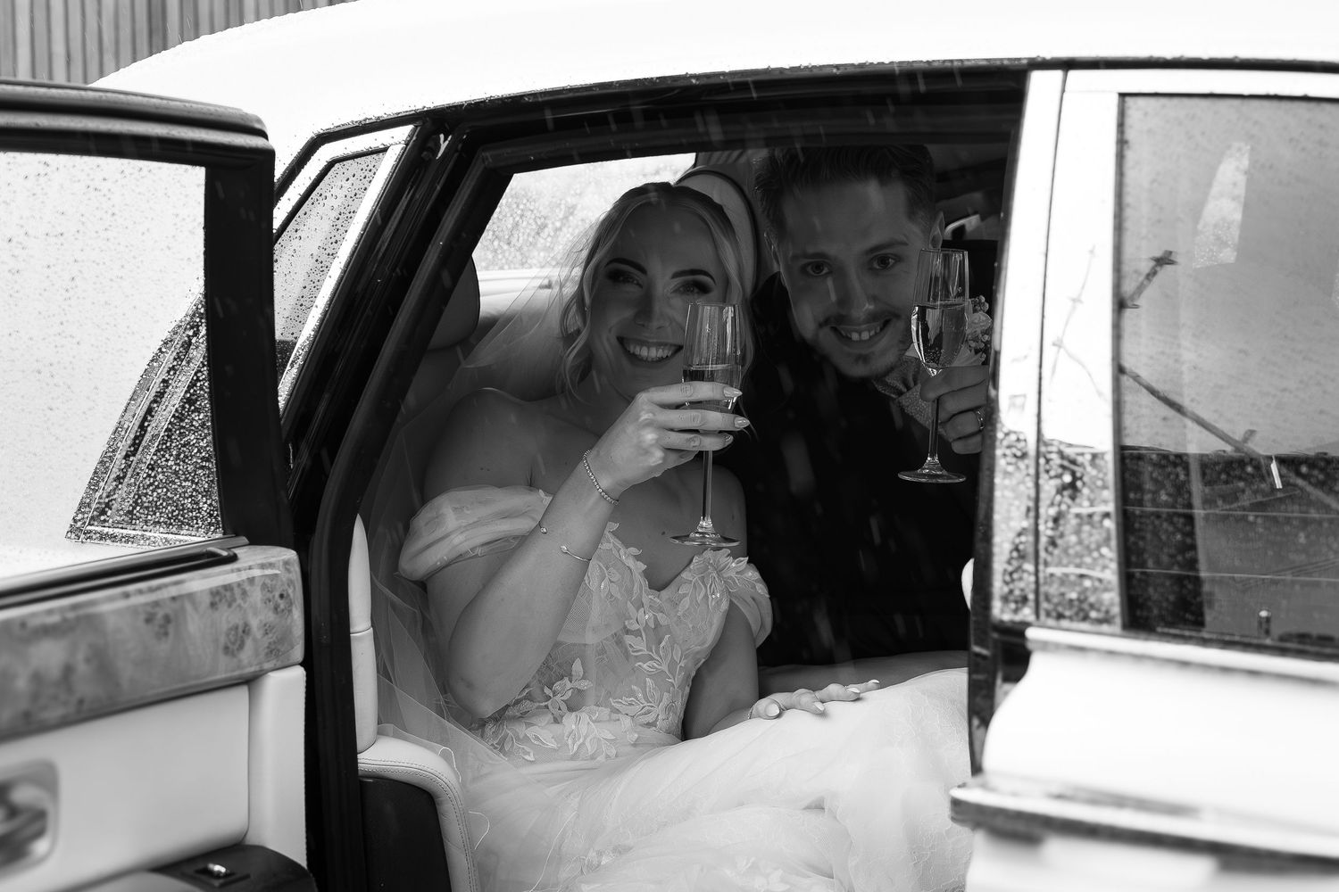 Smiling bride and groom toasting with champagne inside classic white wedding car.
