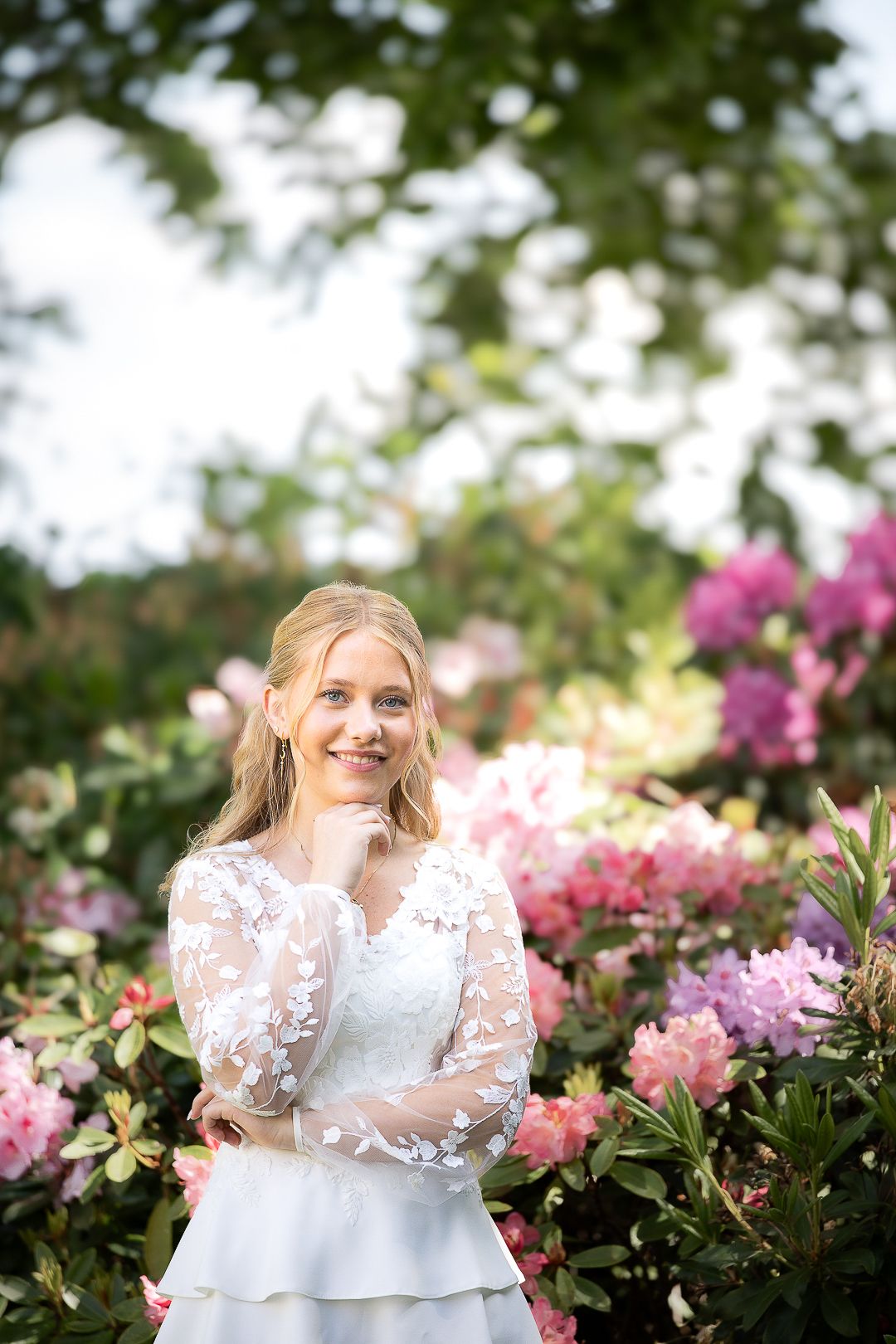 Smiling blonde girl in white lace confirmation dress posing among pink and purple flowers.