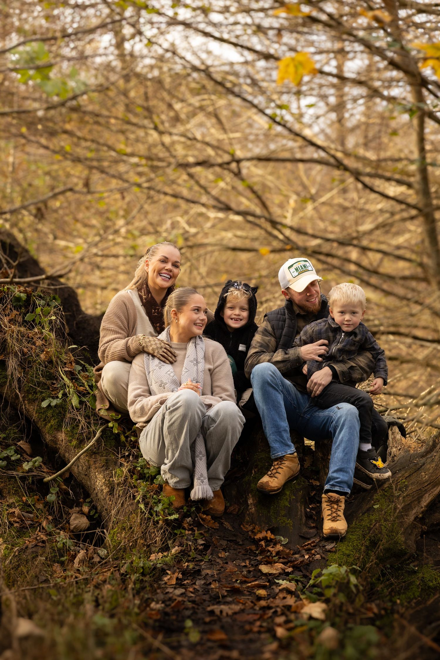 Happy family of five sitting on a fallen log in an autumn forest, surrounded by golden foliage.