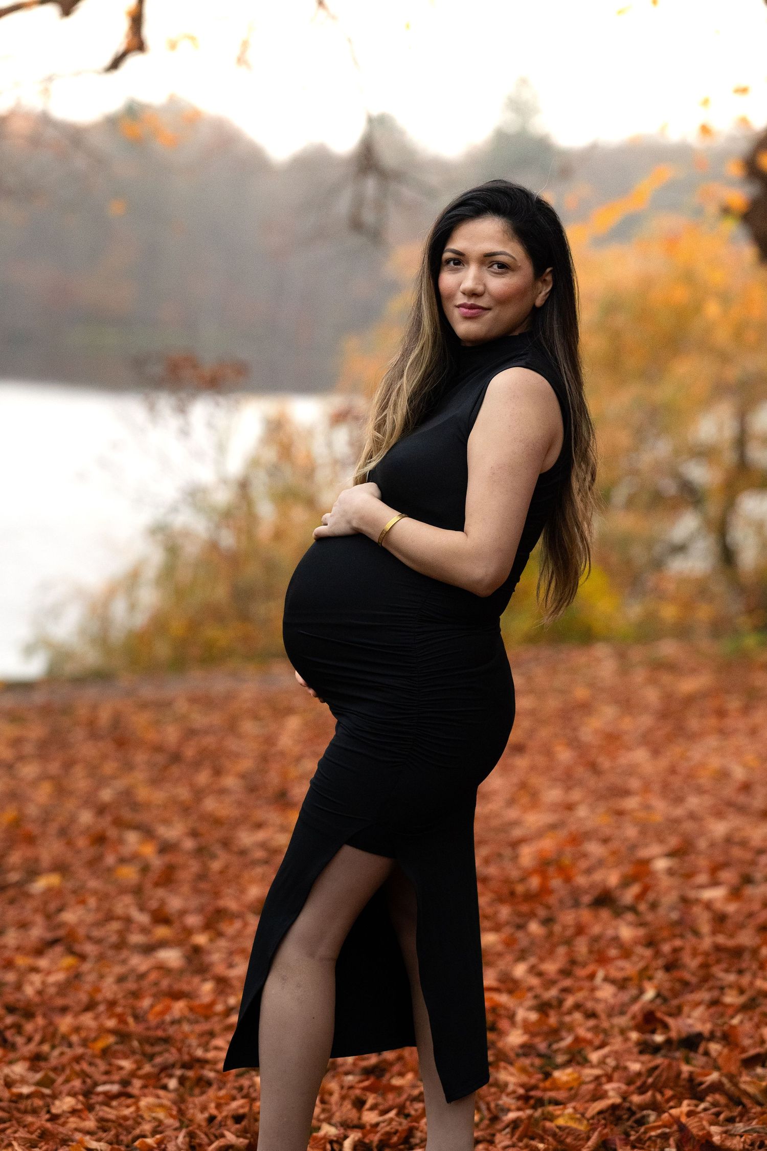 Elegant pregnant woman in fitted black dress holding baby bump among vibrant autumn fallen leaves.