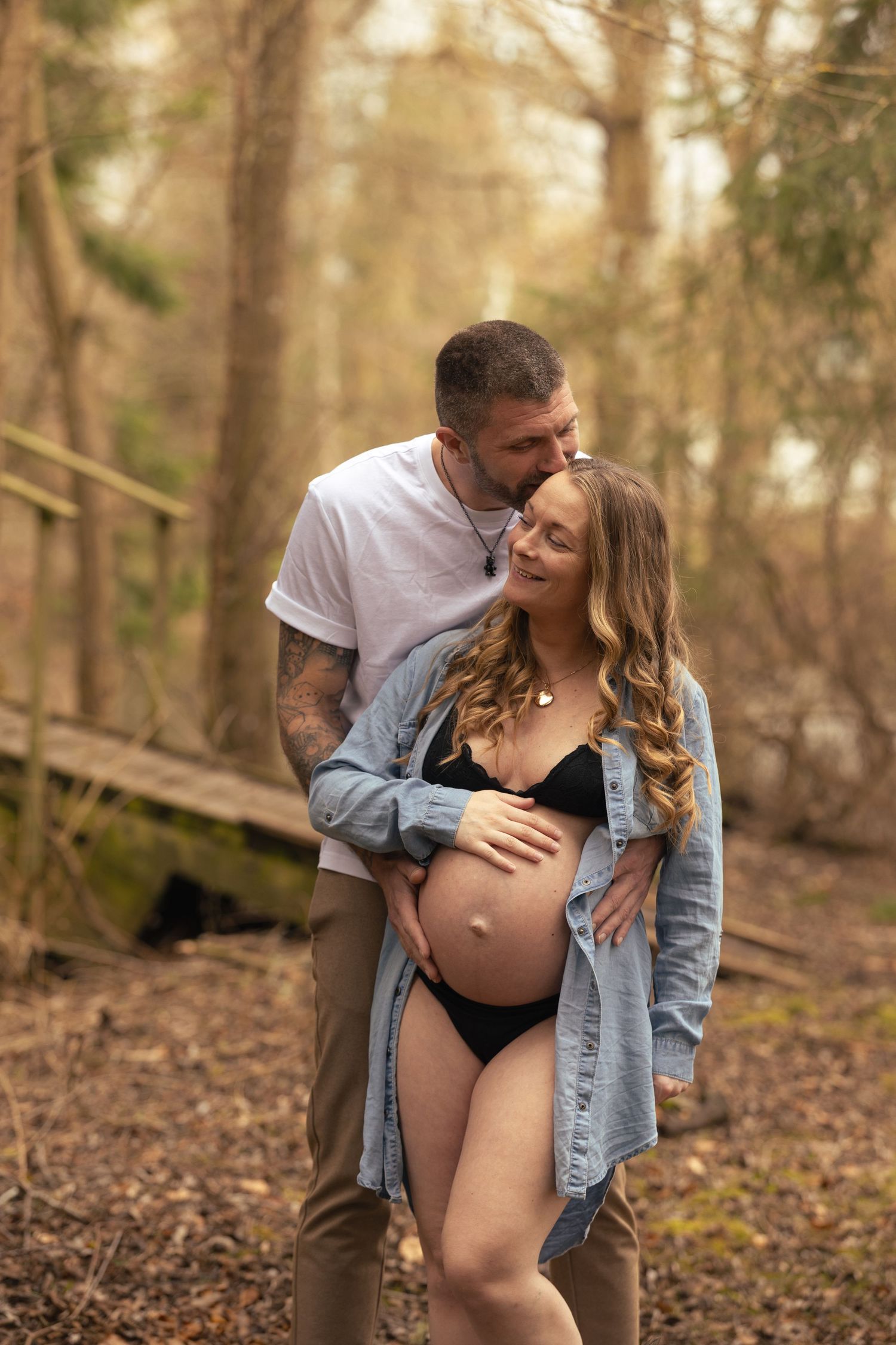 Couple in forest setting, man kissing pregnant woman's head while cradling her bare baby bump.