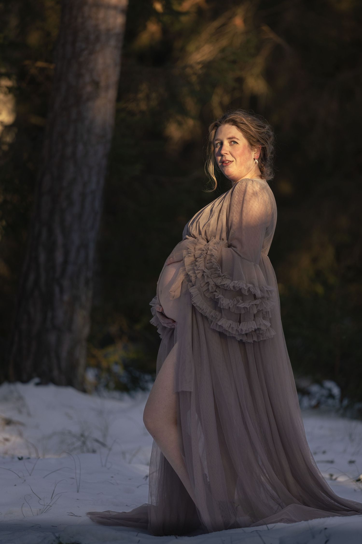 Pregnant woman in elegant taupe chiffon gown posing gracefully in snowy forest during golden hour.