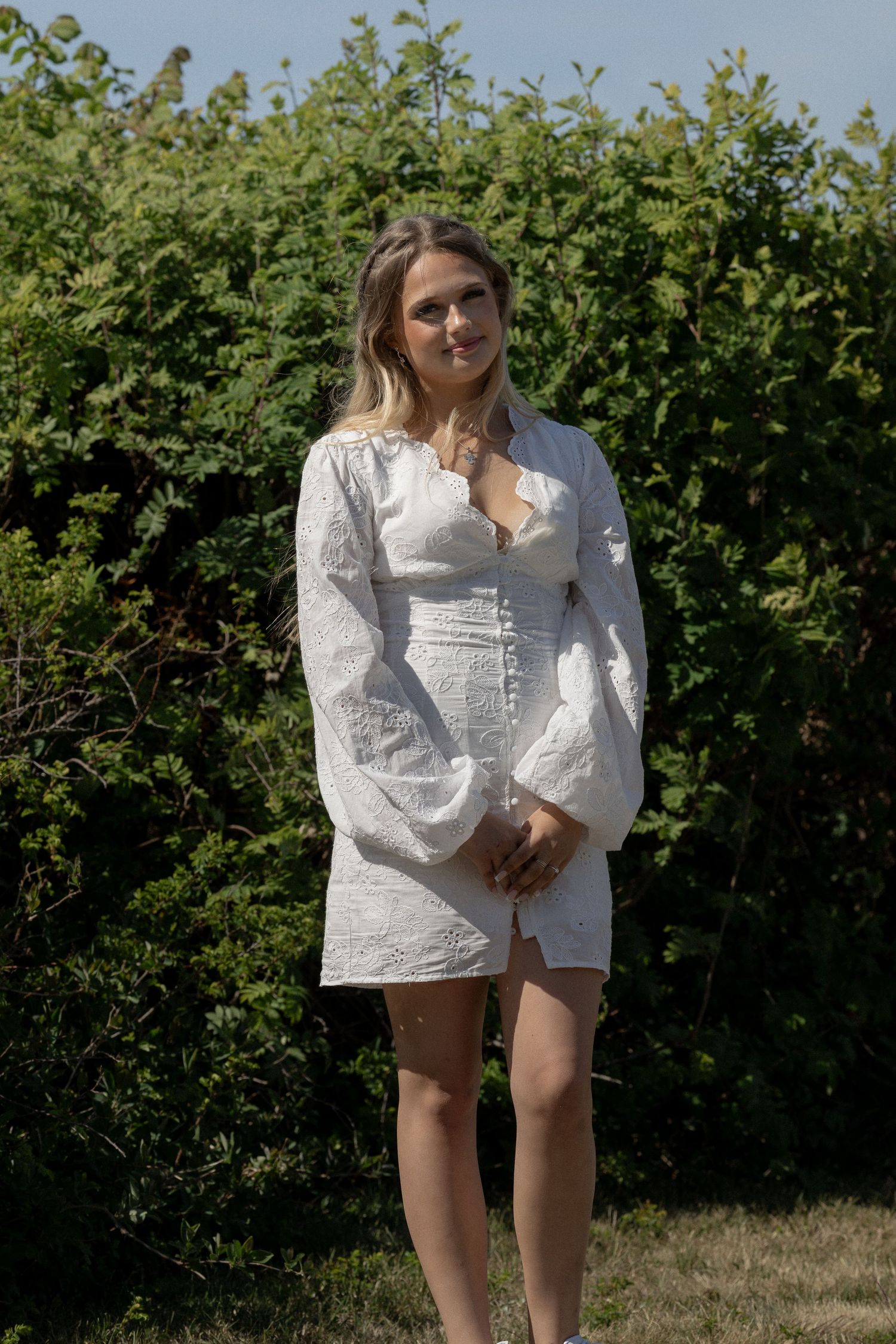 Woman in white lace long-sleeve mini dress standing outdoors in front of green hedges on a sunny day.