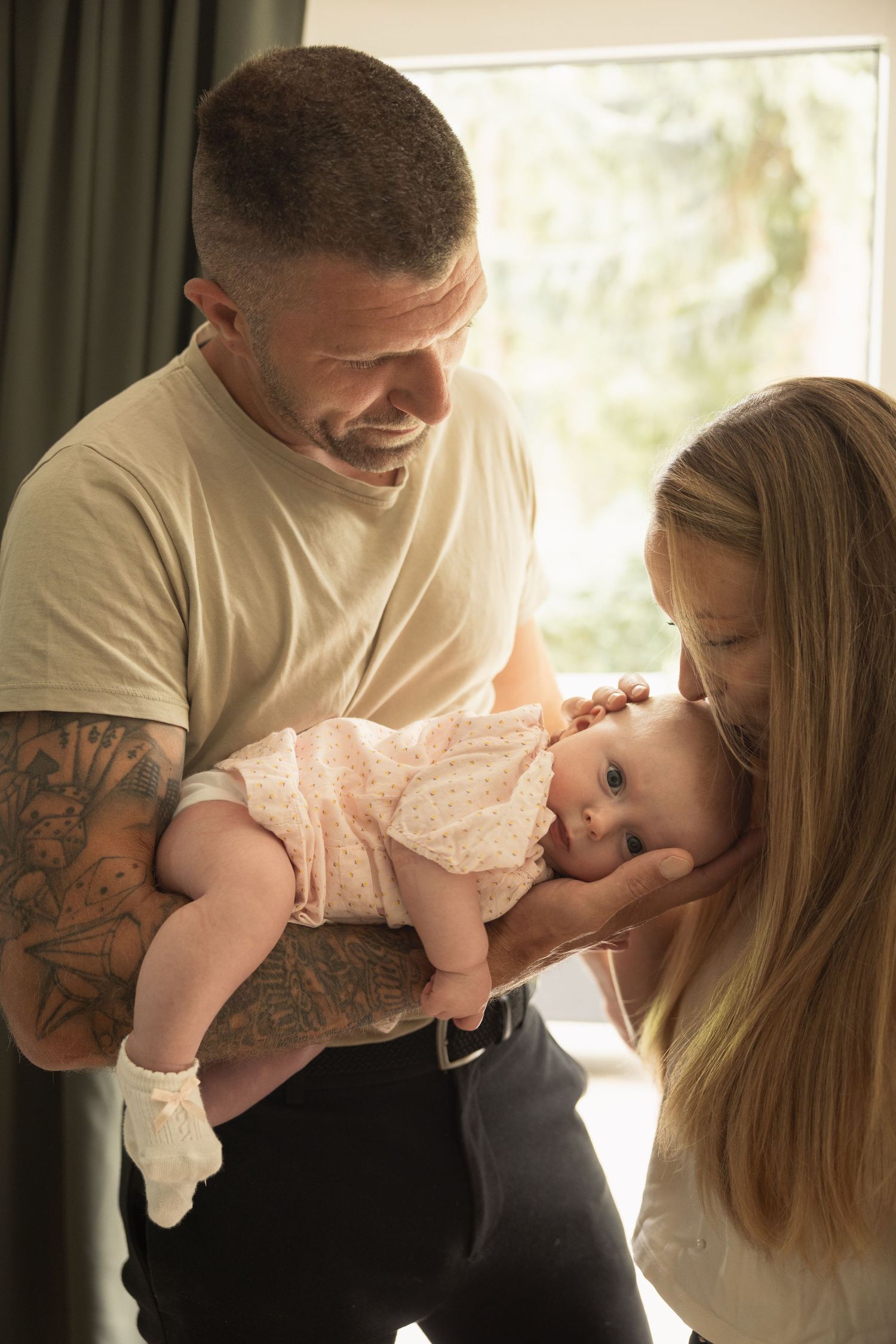 A smiling tattooed father holds his newborn baby girl in a pink outfit while the mother gently kisses her cheek.