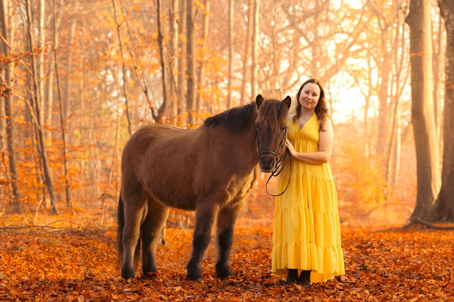 Woman in yellow dress standing with brown horse in autumn forest with golden foliage.