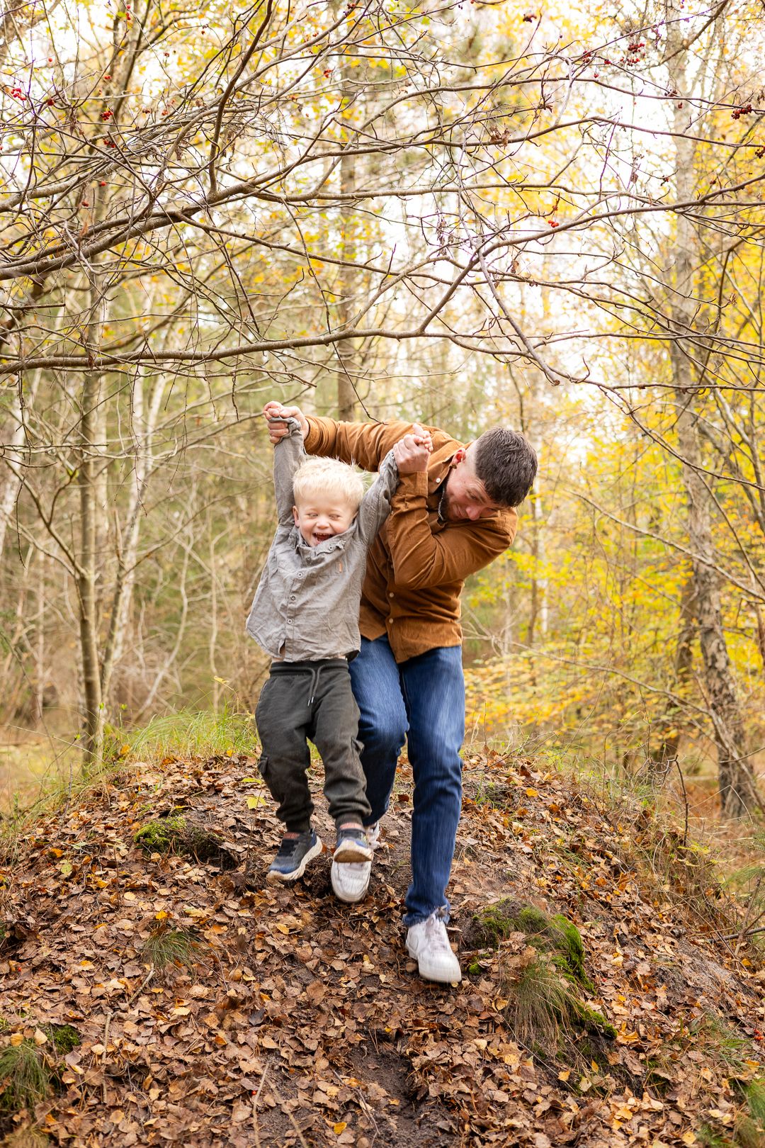 Father and son laughing and playing together on a leaf-covered hill in an autumn forest.