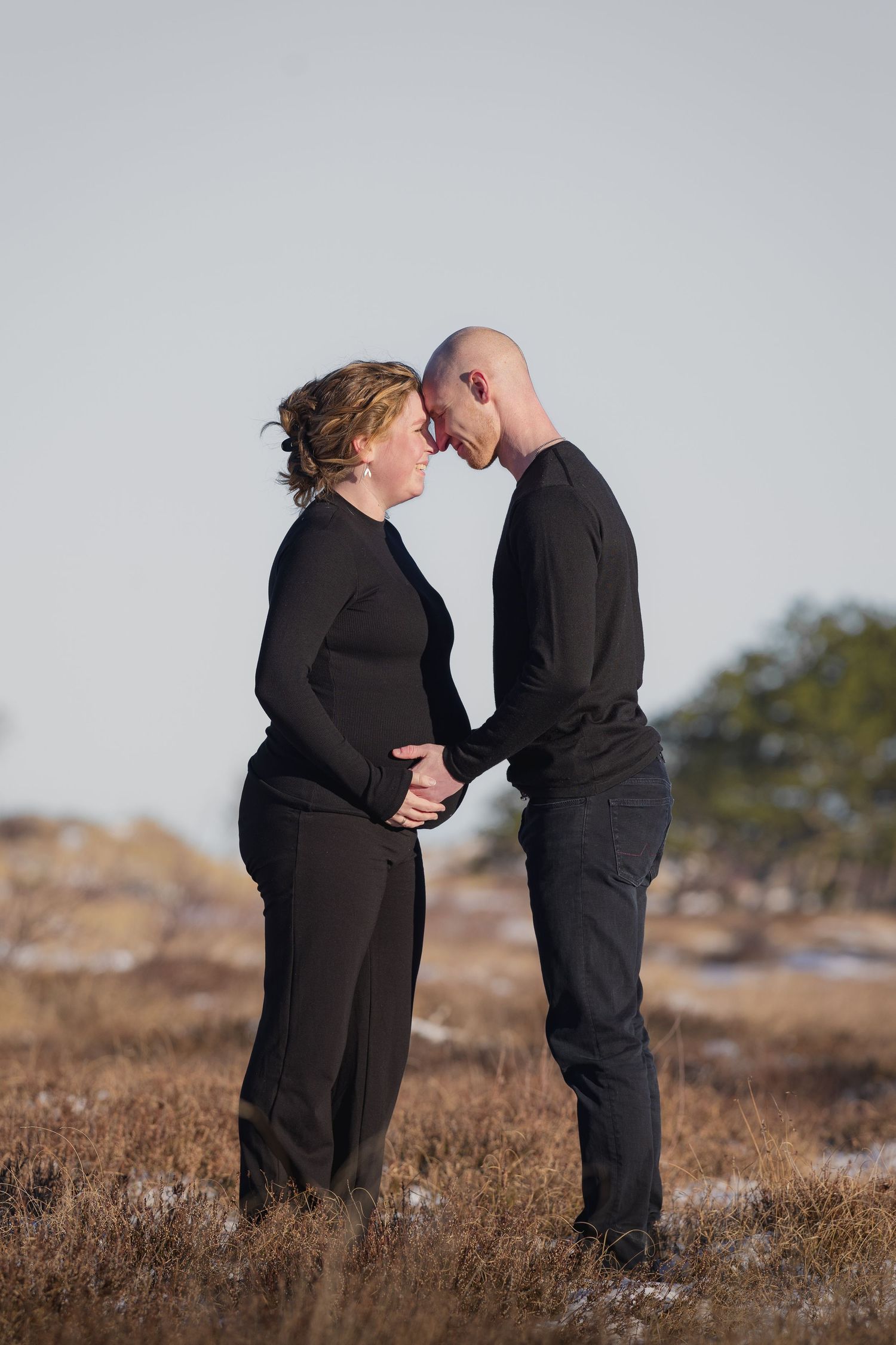 Expectant couple in matching black outfits touching foreheads tenderly in a winter landscape.