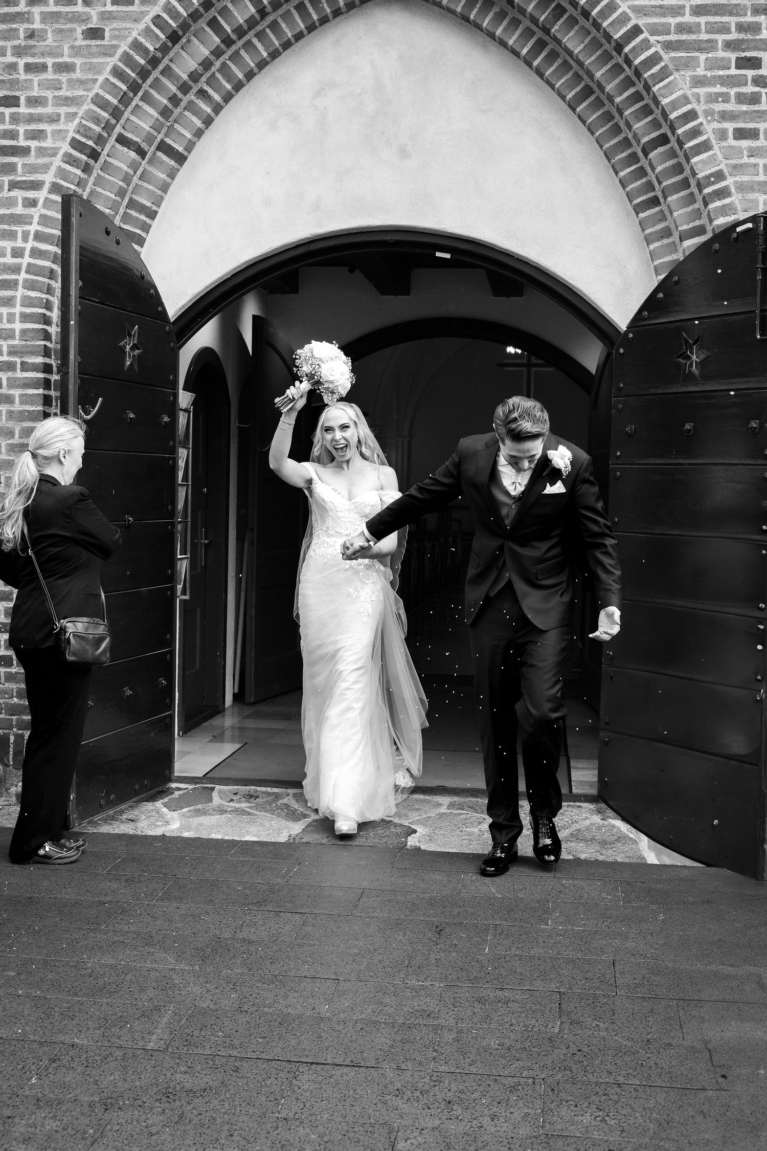 Black and white photo of newlyweds exiting church as bride raises bouquet in celebration.