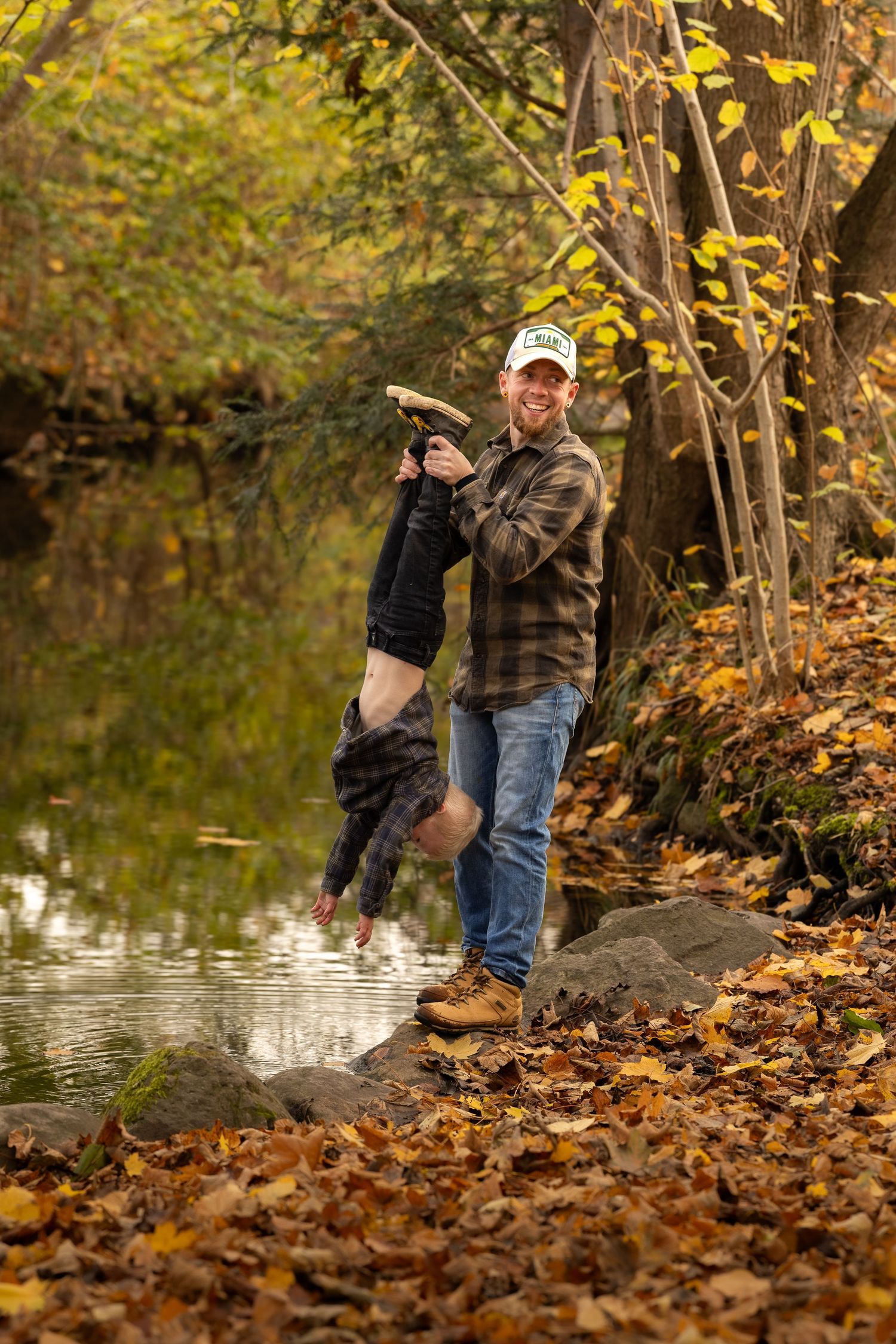 Laughing father playfully holding his child upside down by a reflective pond in autumn woods.