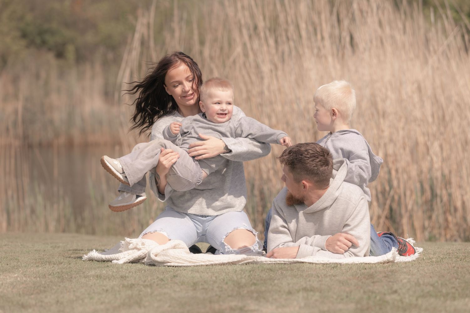 Joyful family sitting on a blanket outdoors, mother lifting baby while father and toddler play nearby.