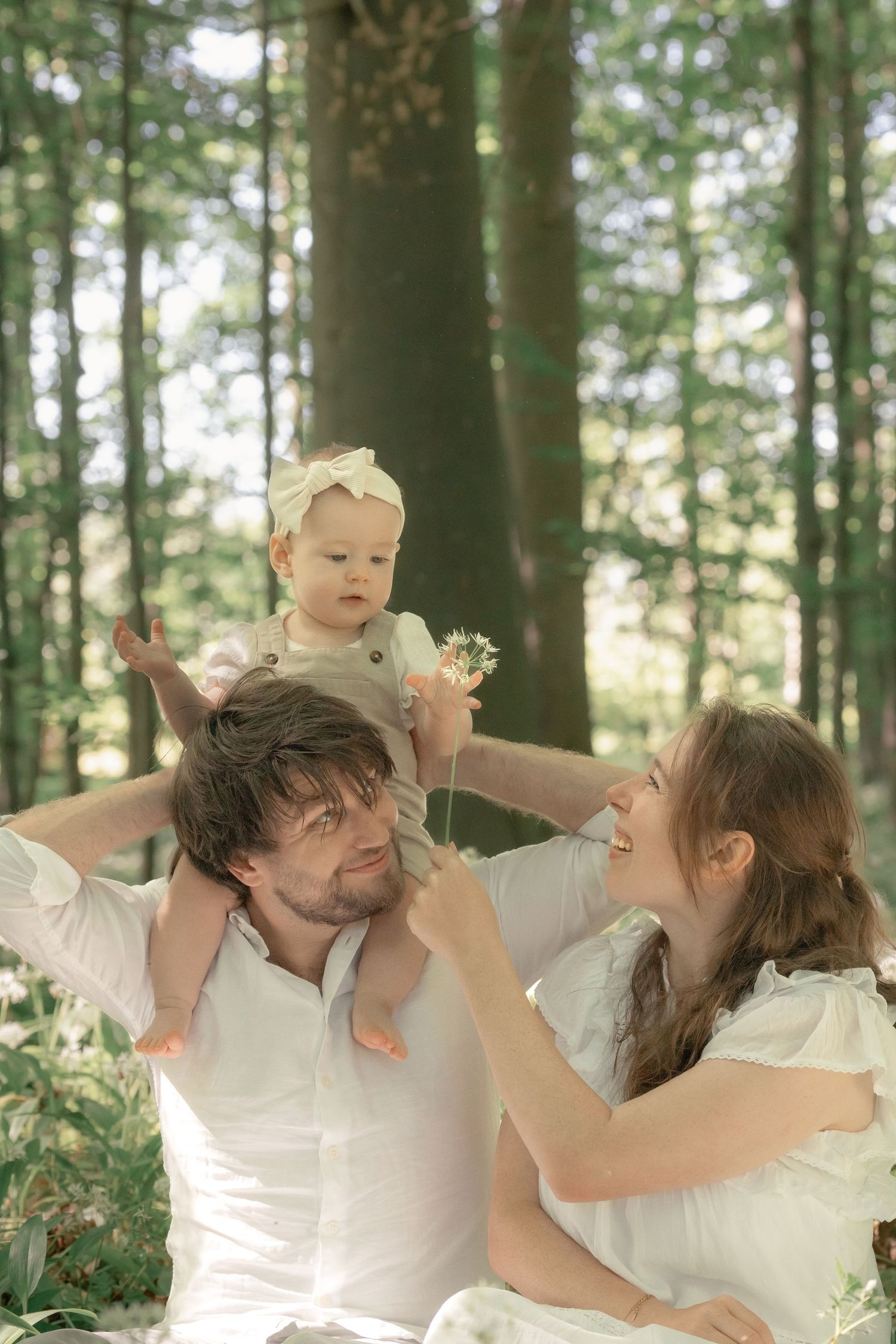 Family photo session in forest with father holding baby on shoulders while mother smiles nearby.