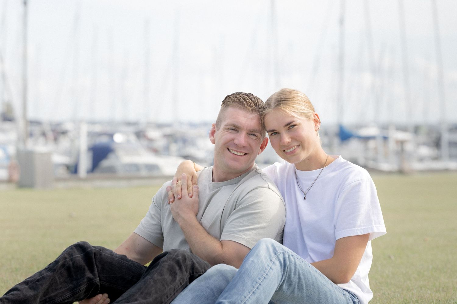 A smiling young couple sits together on green grass with a marina full of sailboats in the background.