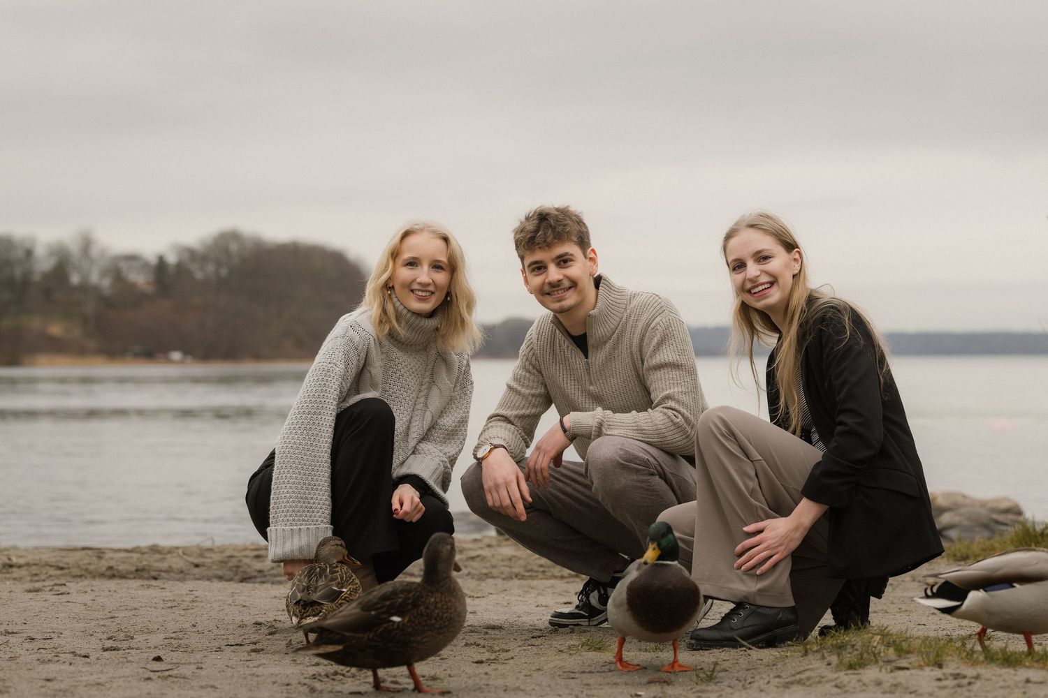 Three young adults crouch on a rocky beach near ducks by a calm lake on an overcast day.