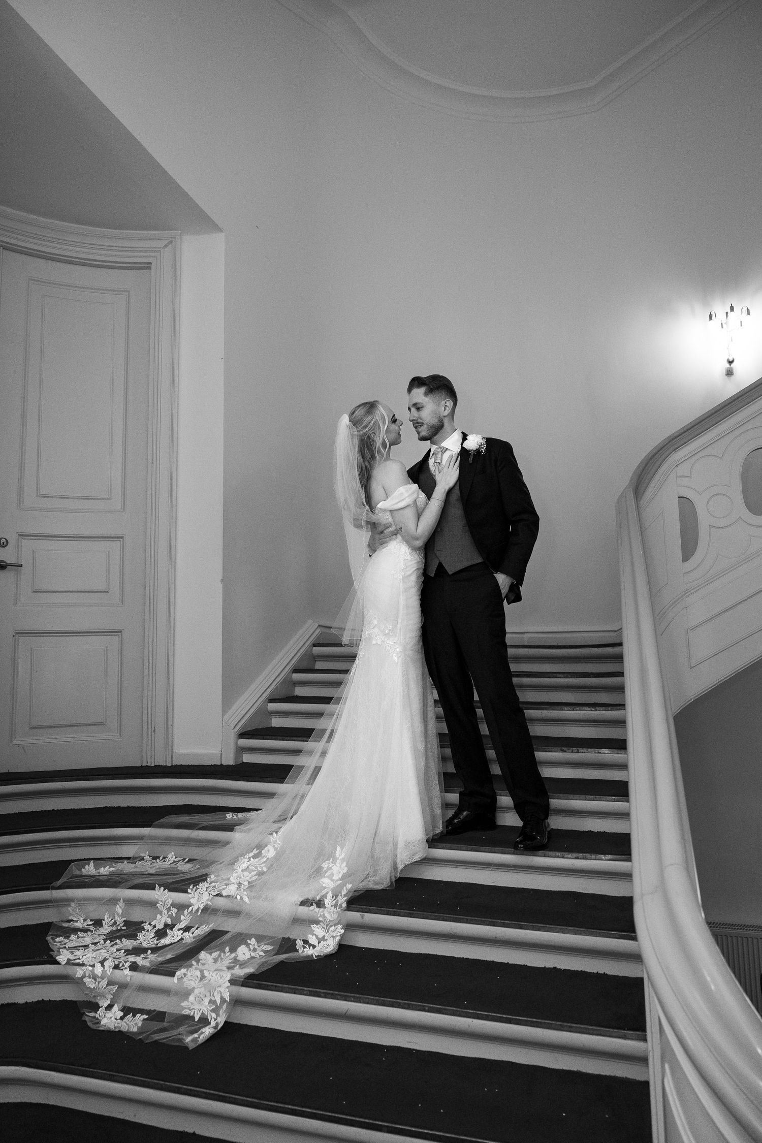 Bride and groom share intimate moment on sweeping grand staircase in stunning black and white photo.