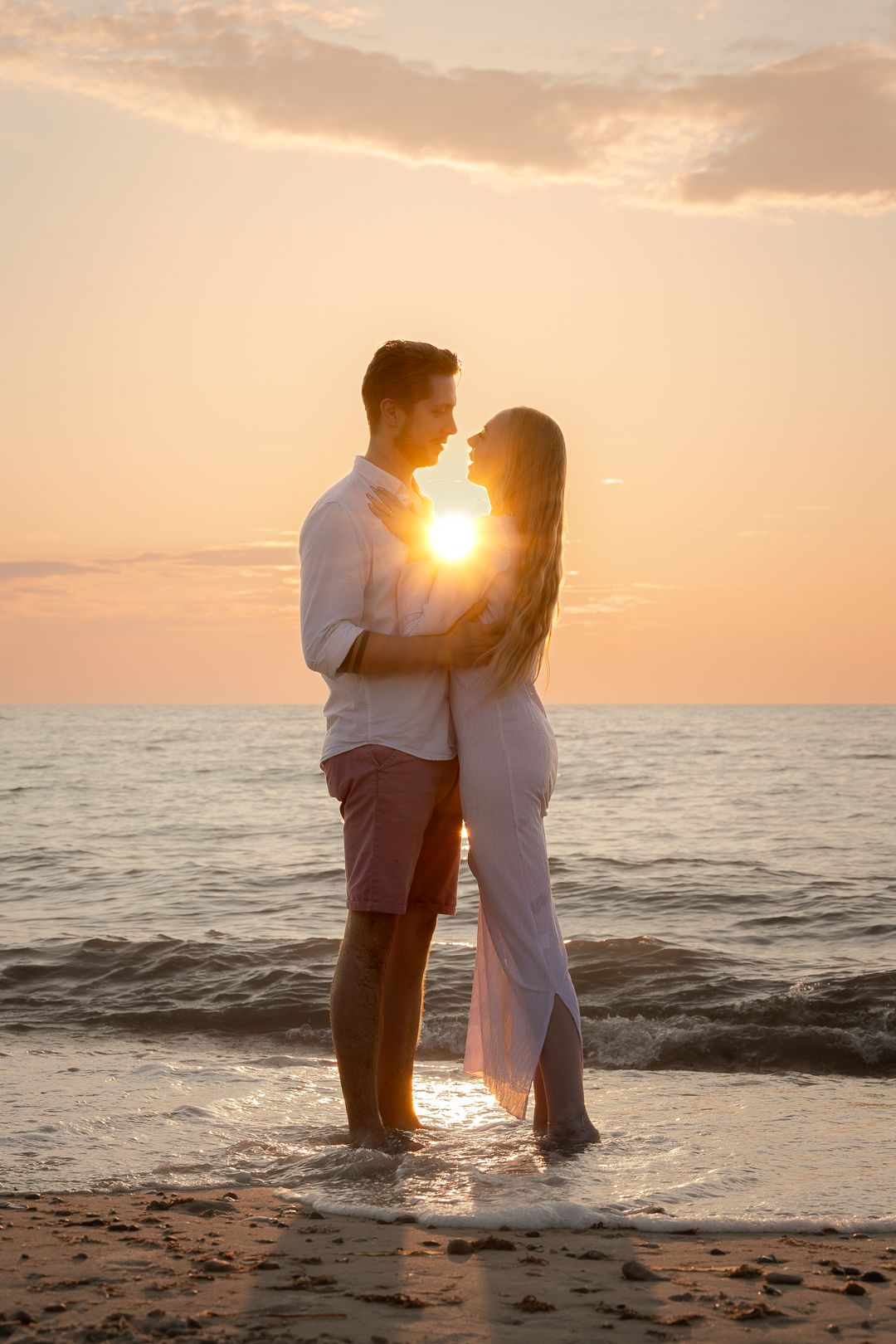 A romantic couple embraces while standing in shallow ocean waves, backlit by a stunning golden sunset.