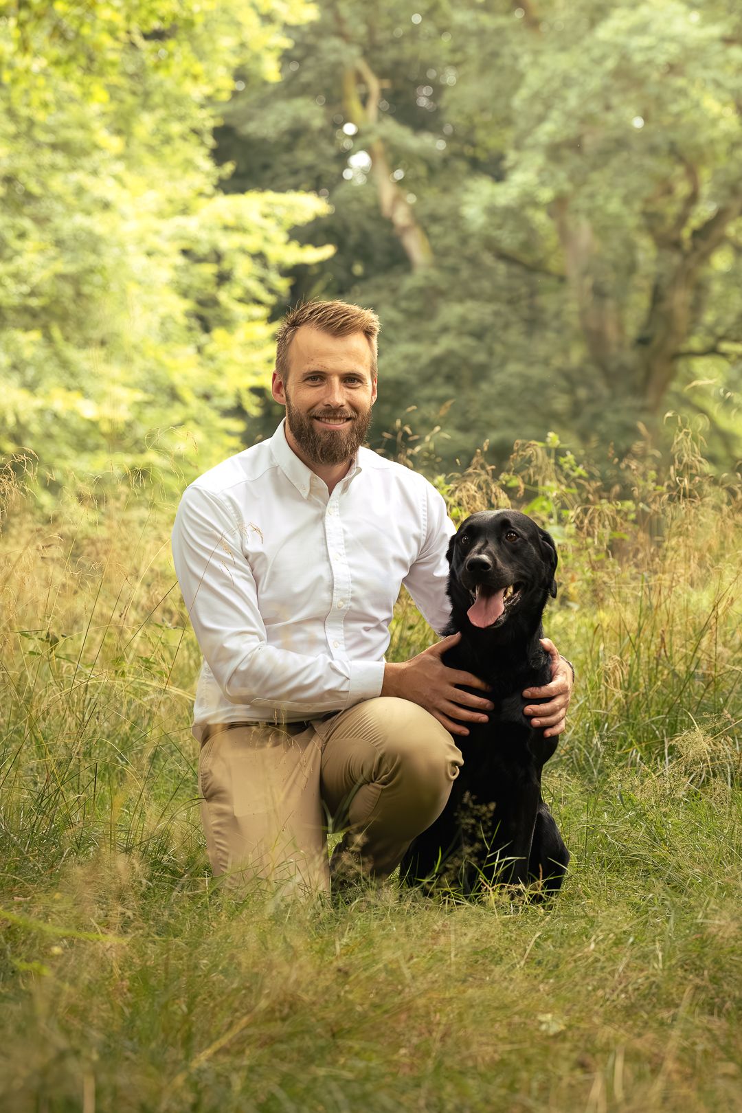 Smiling bearded man in white shirt posing with black Labrador dog in lush green meadow.