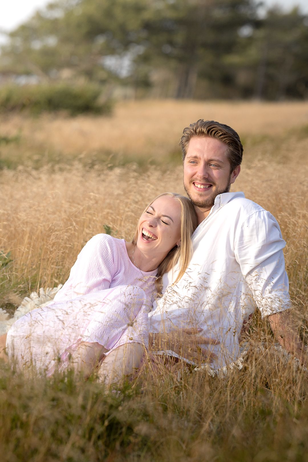 A laughing couple sits together in a golden field of tall grass, enjoying a sunny outdoor moment.