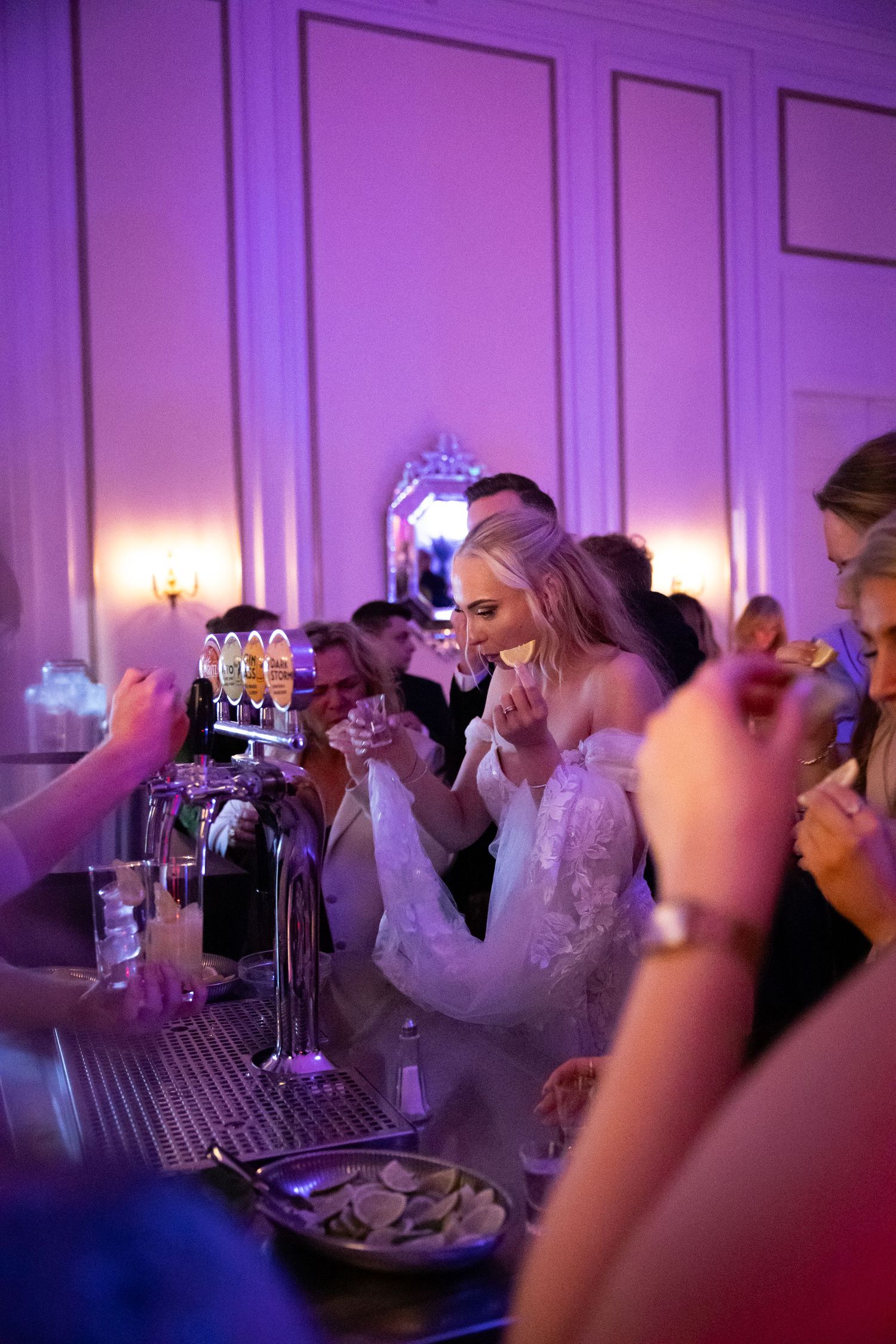 Bride in a lace wedding gown laughing at the bar with guests under purple lighting at her wedding reception.
