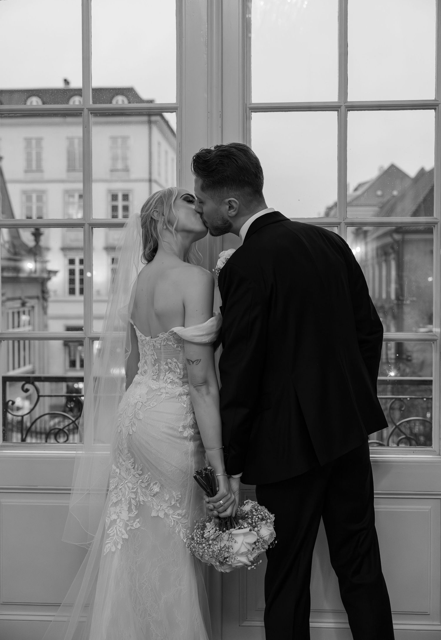 Bride and groom sharing romantic kiss by large window in black and white.
