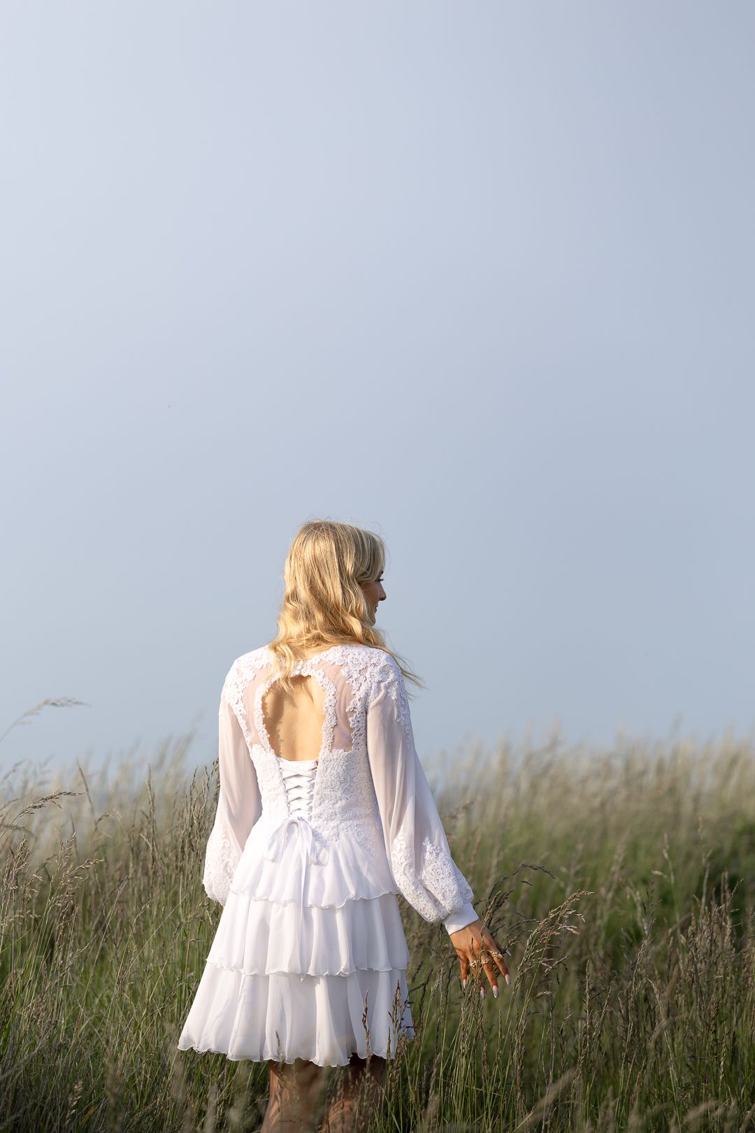 Back view of blonde woman in white lace corset dress walking through tall grass field.