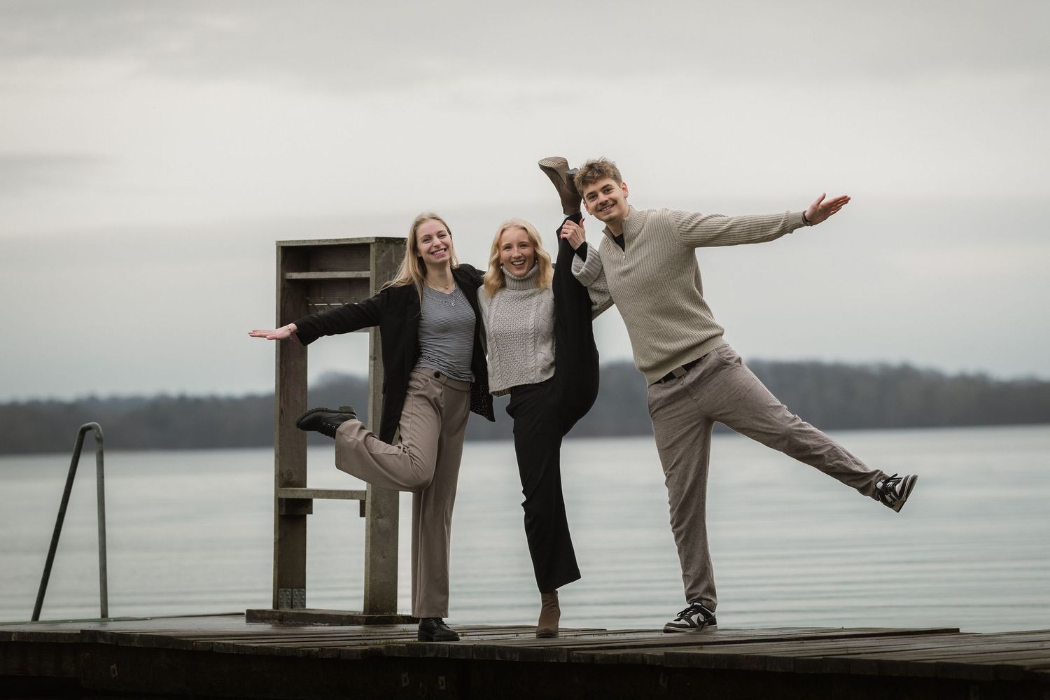 Three young adults playfully pose on a wooden dock beside a calm lake on an overcast day.