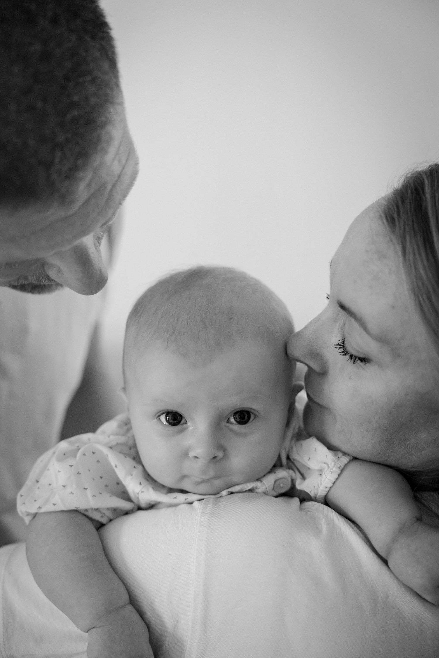 Black and white portrait of parents kissing their newborn baby on cheeks indoors.