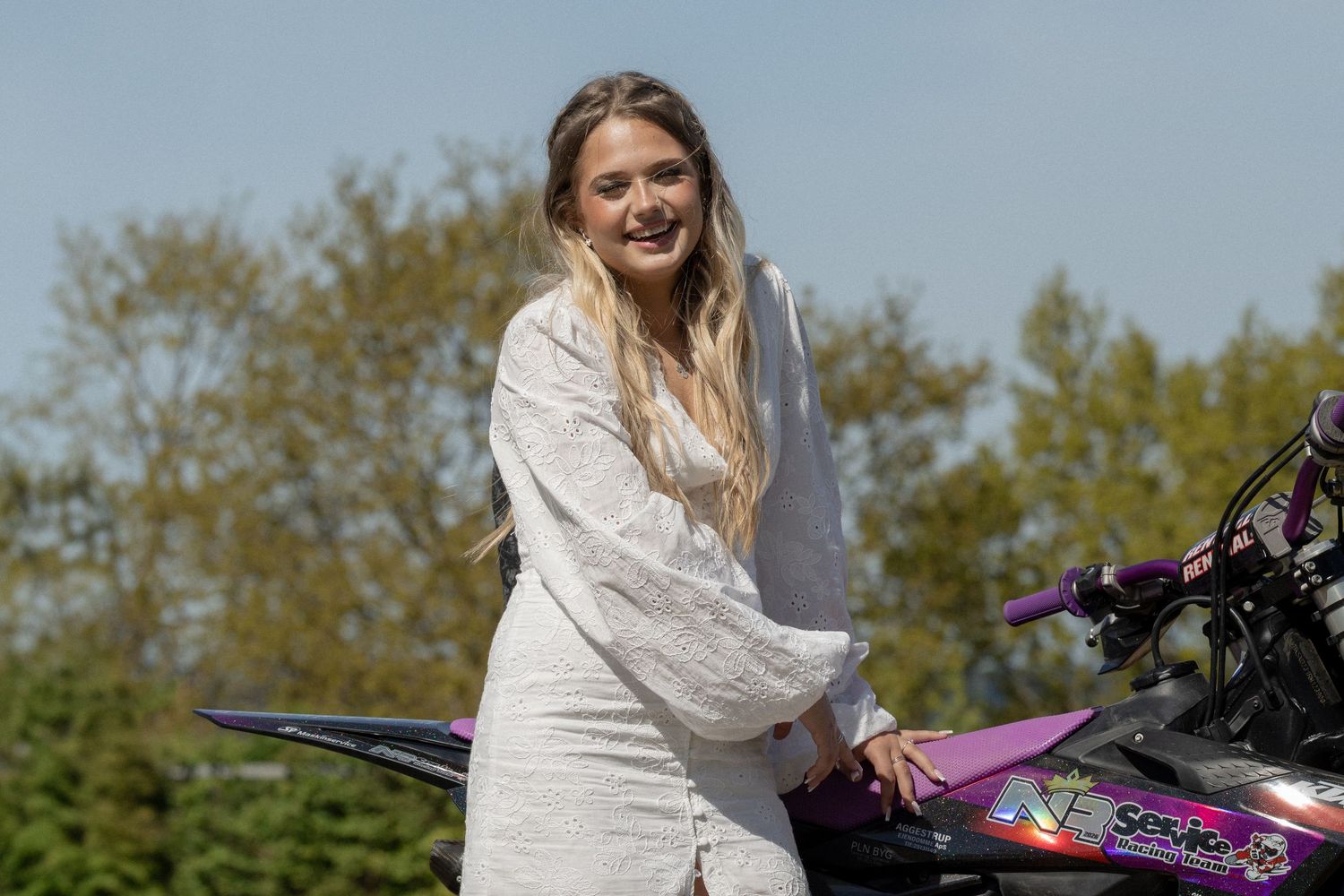 Young blonde woman in white dress smiling beside a purple motorbike outdoors with trees in background.