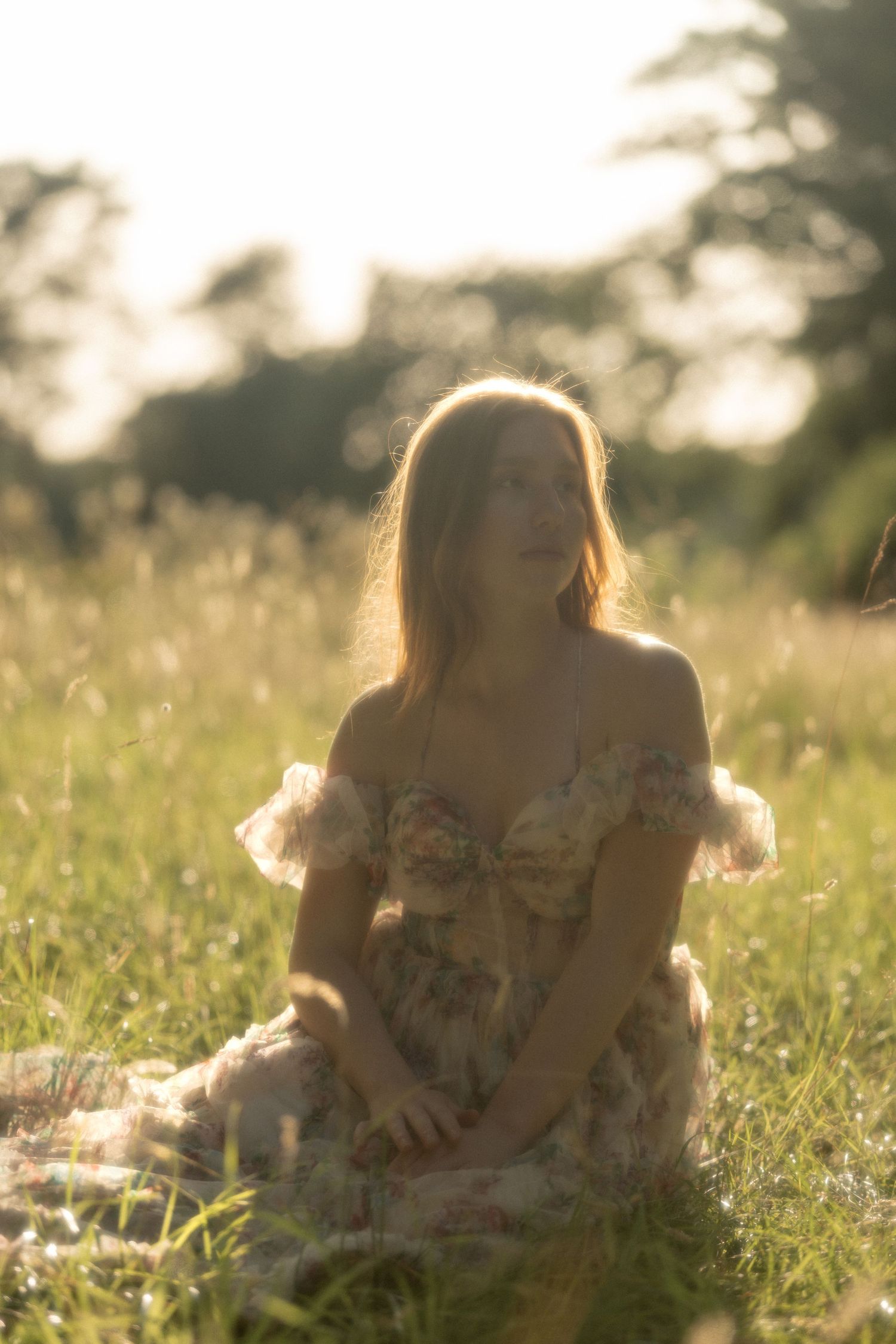 Young woman in floral off-shoulder dress sitting in sunlit meadow grass, golden hour backlight.