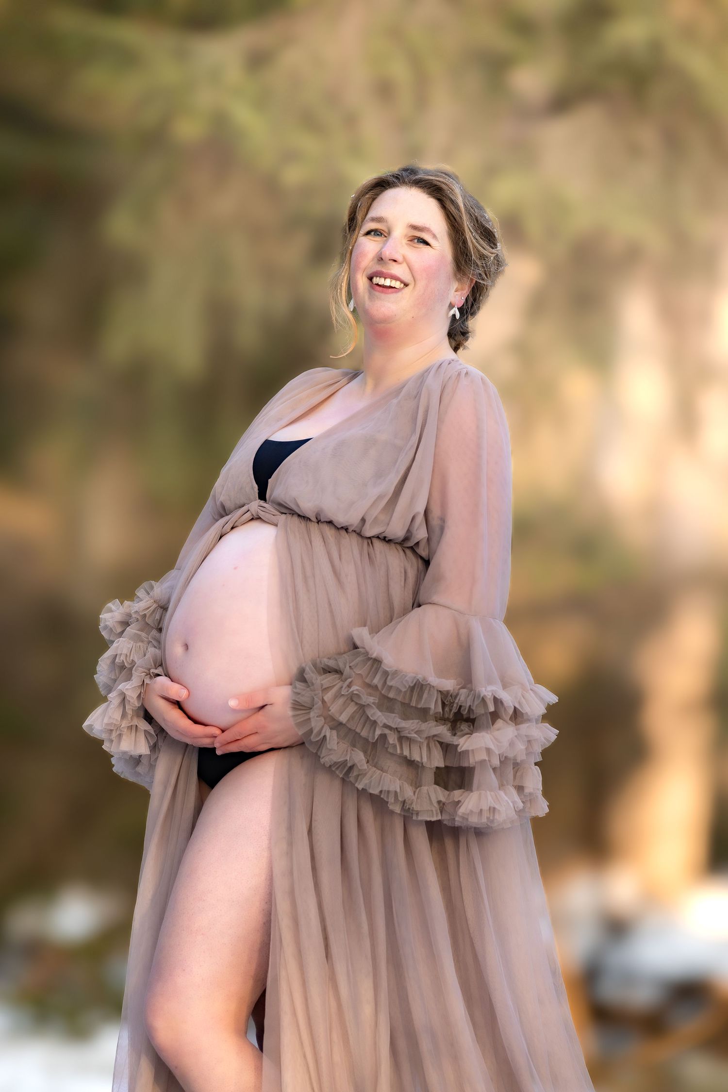 Smiling pregnant woman in sheer taupe robe holding baby bump outdoors in warm natural light.