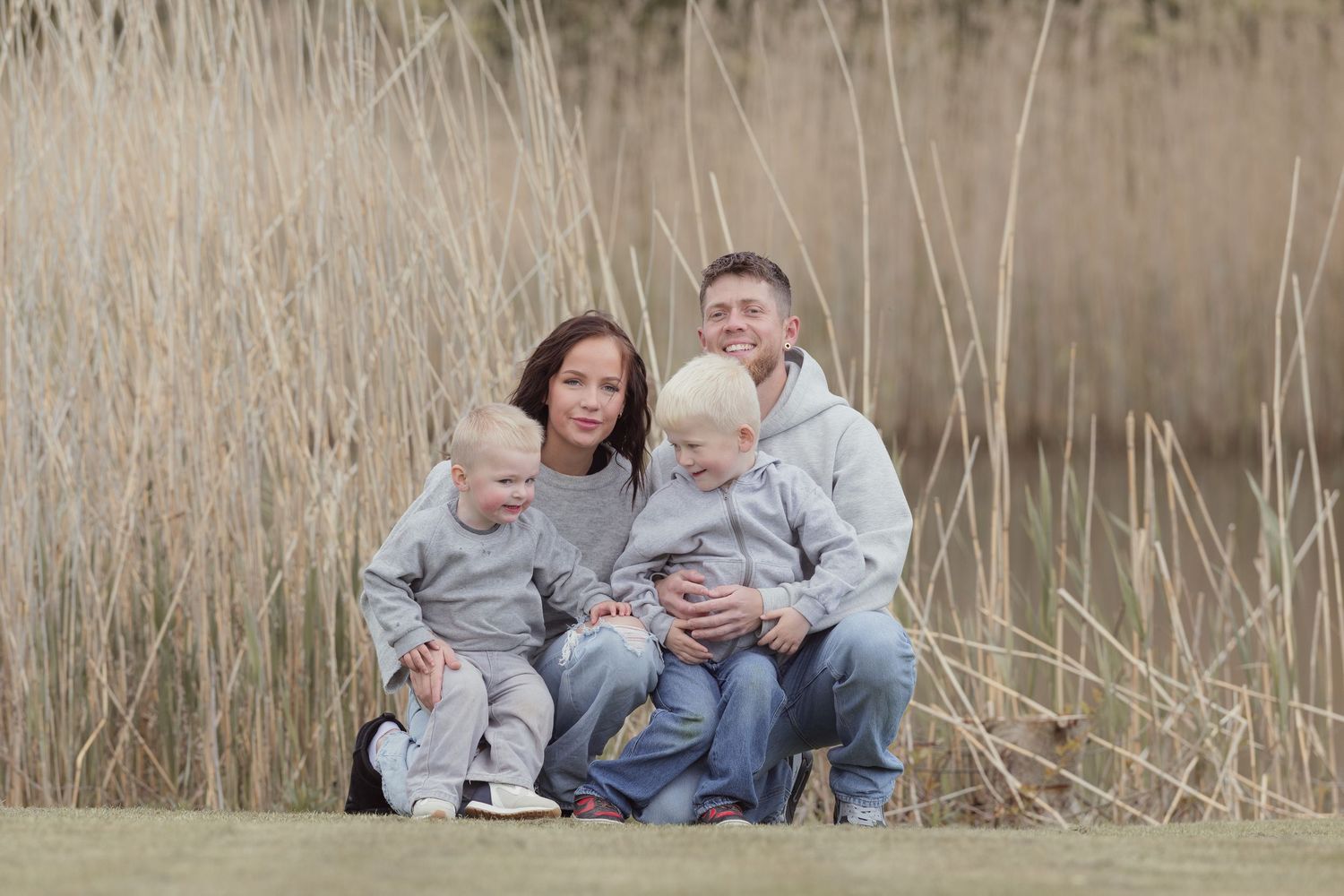 Happy family of four in matching grey hoodies sitting together in front of tall golden reeds.