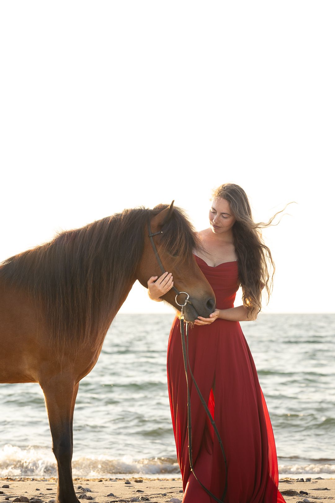 Woman in red dress gently stroking brown horse face on beach with ocean behind.