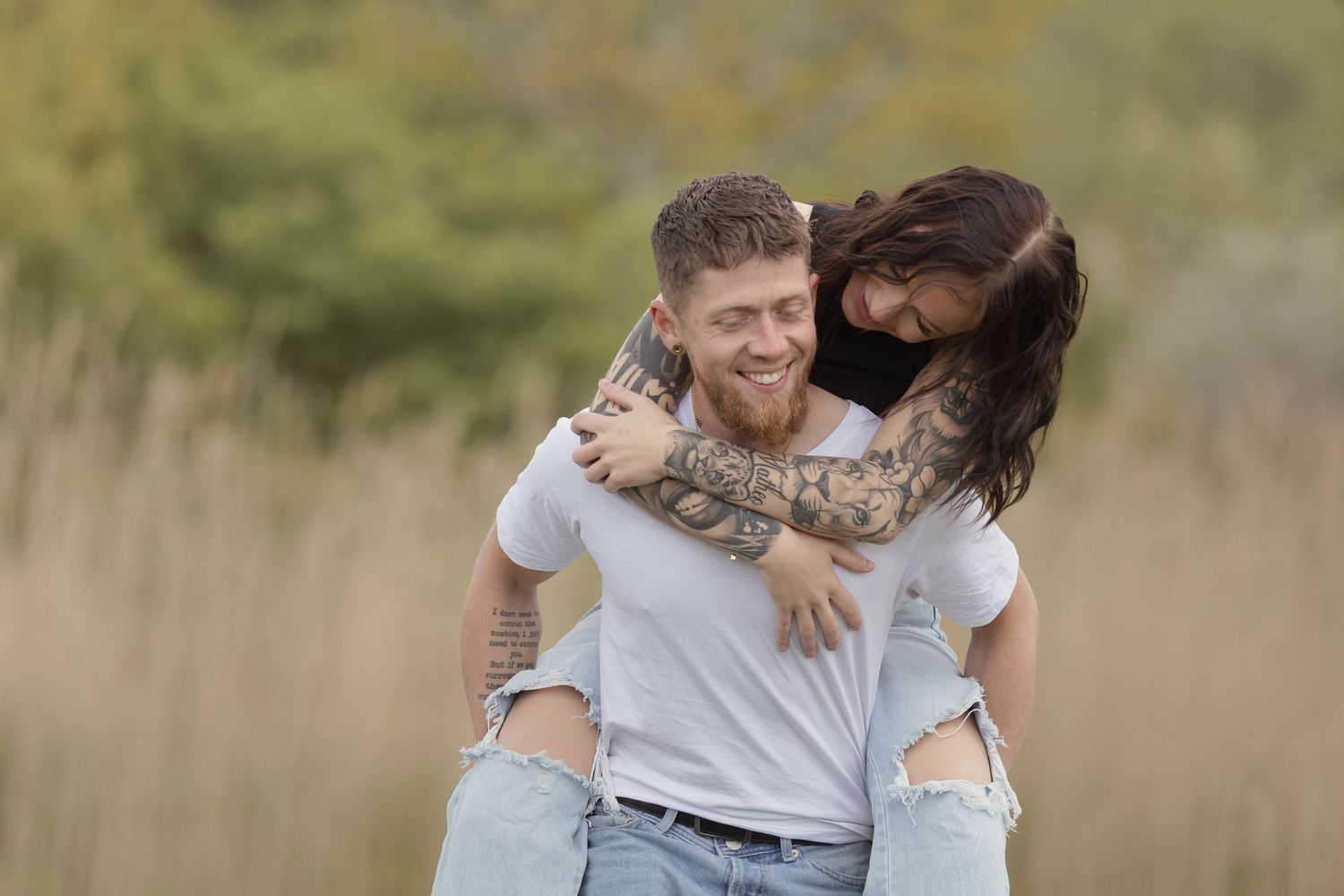 Smiling man with tattoos giving a woman a piggyback ride in a golden field outdoors.