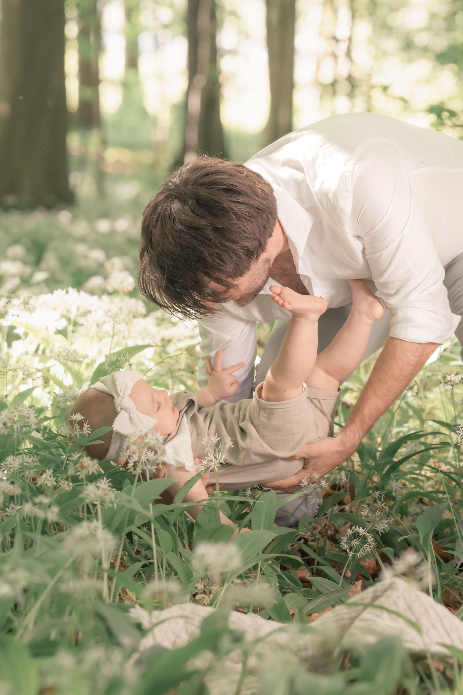 A loving father leans down to interact with his baby lying among white forest flowers.