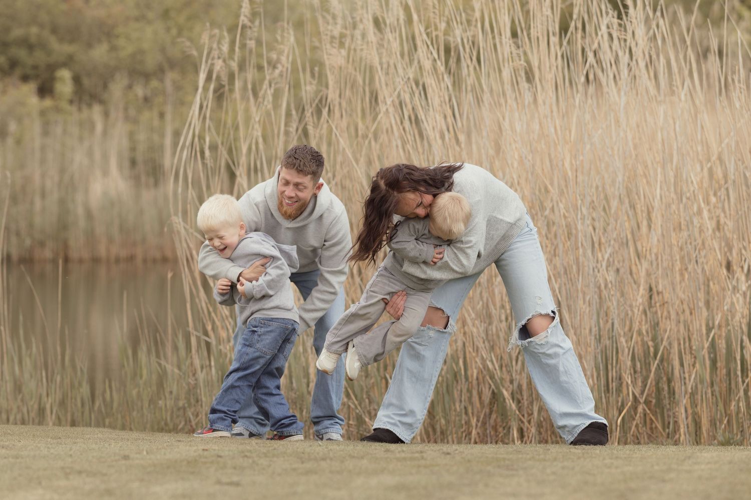 Playful family moment outdoors as parents lift laughing children near a reed-lined lake.