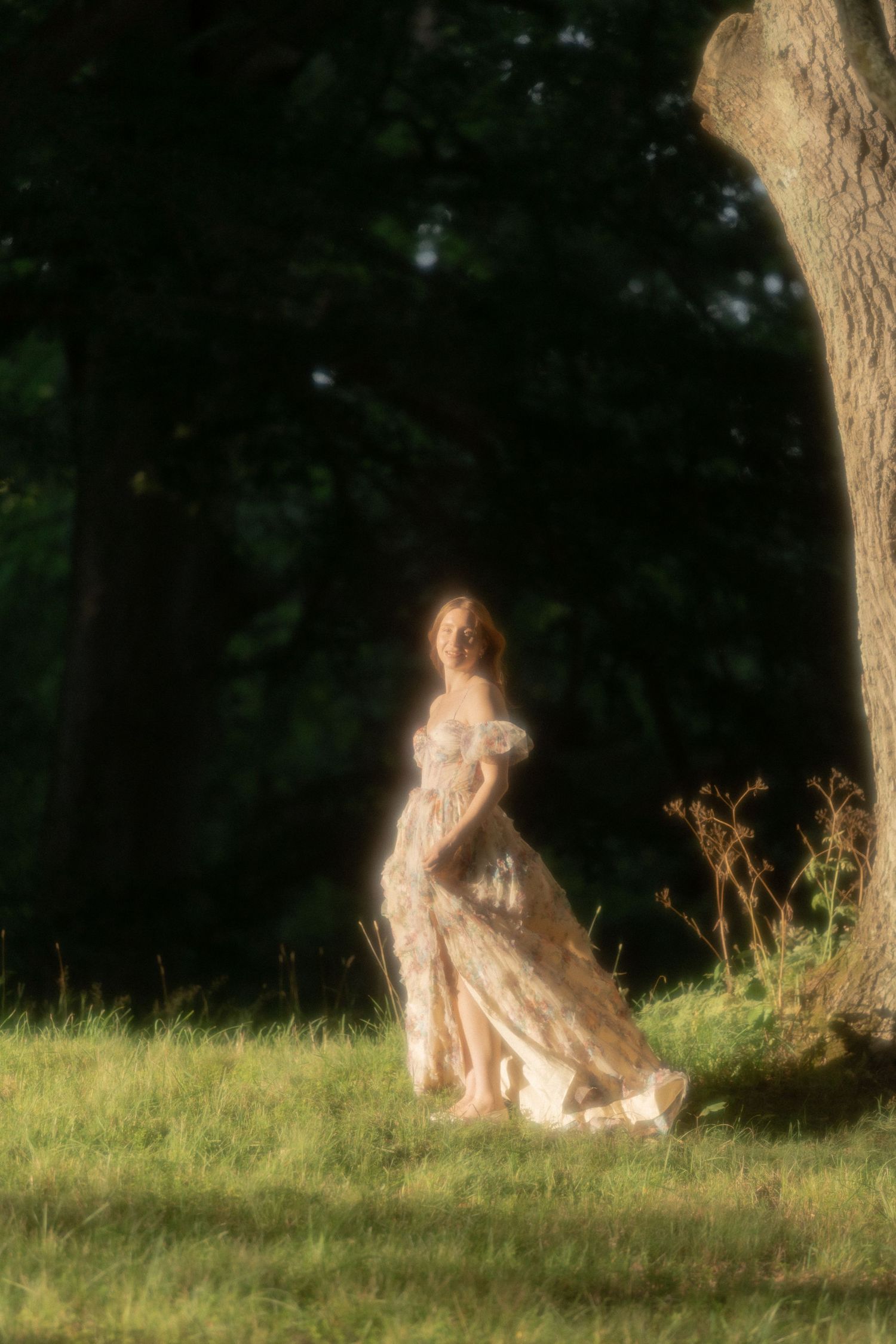 Woman in floral gown standing in sunlit forest clearing beside a large tree trunk.