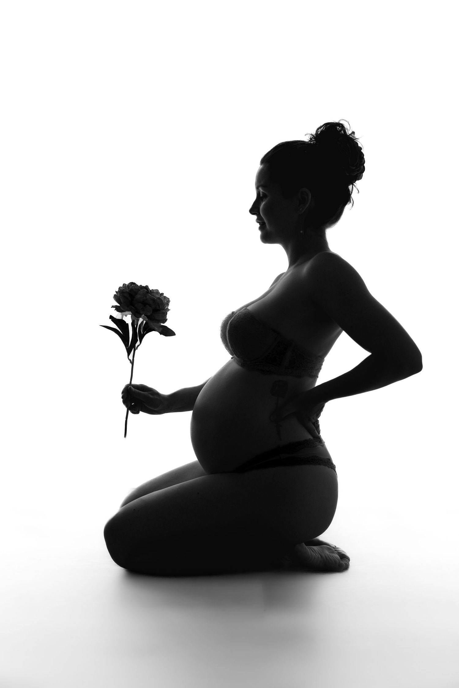 Black and white silhouette of a pregnant woman kneeling, holding a flower, showcasing a beautiful maternity portrait.