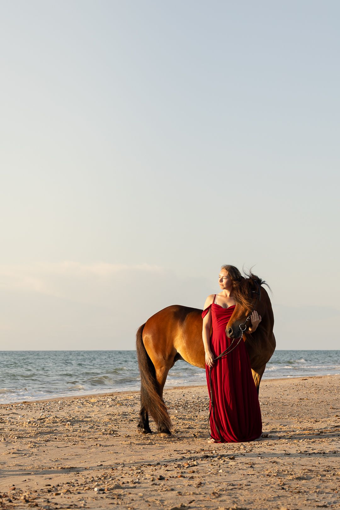 Woman in flowing red dress standing beside brown horse on sandy beach at golden sunset.