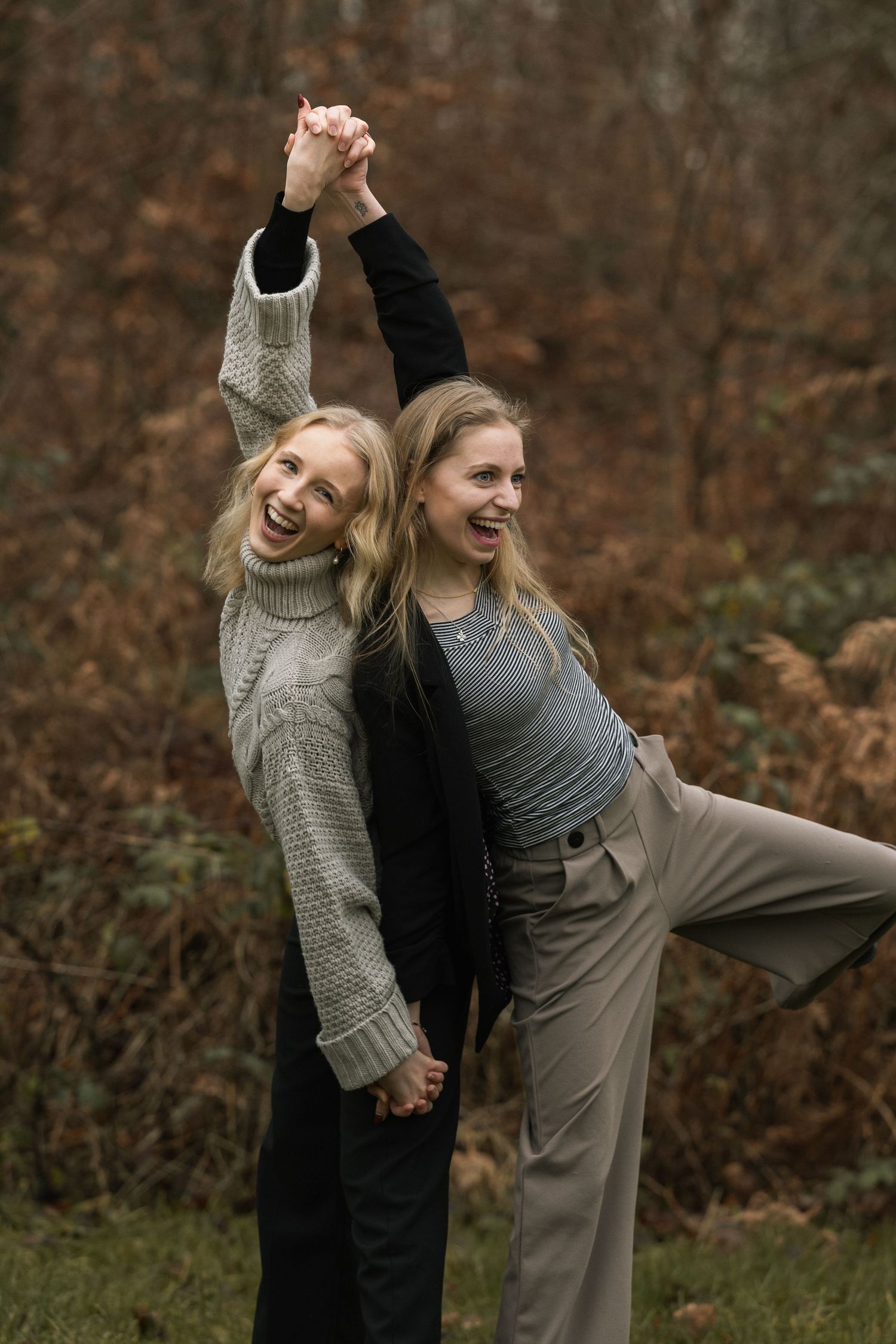 Two laughing blonde women holding hands with arms raised in a playful pose in an autumn forest.