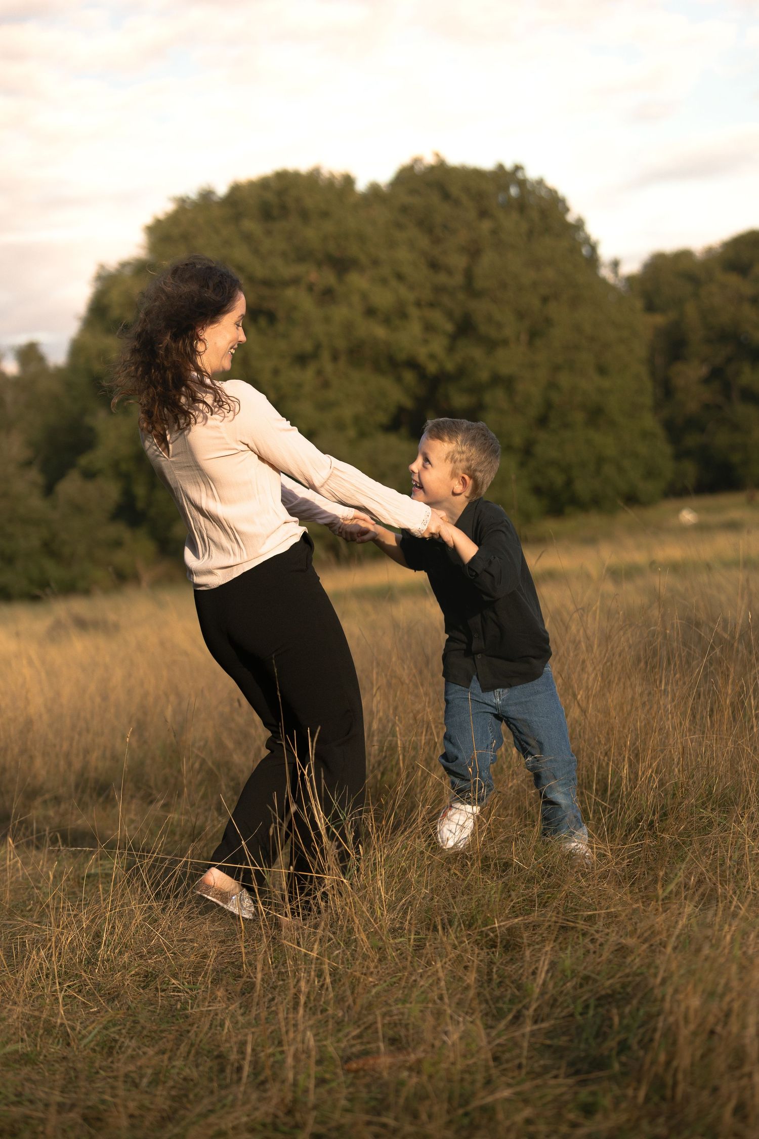 Mother and young son holding hands spinning playfully in golden grass field at sunset.