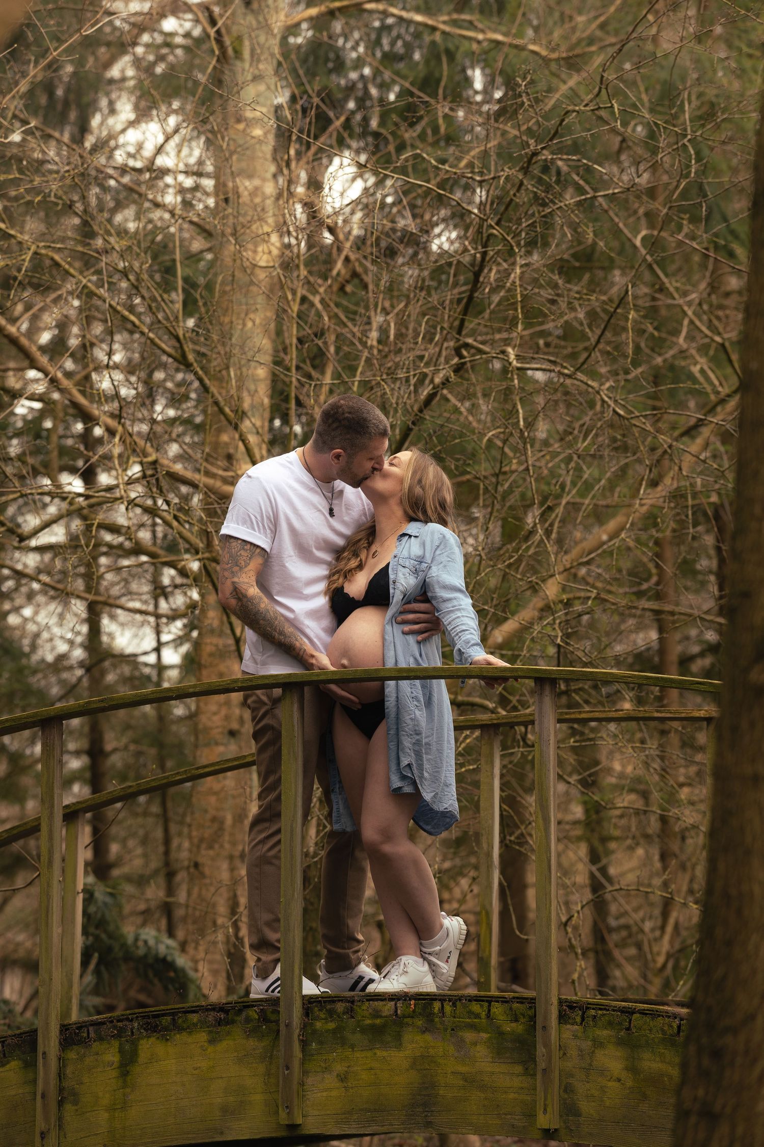 Expecting couple sharing a kiss on moss-covered bridge surrounded by tall bare trees in woodland.