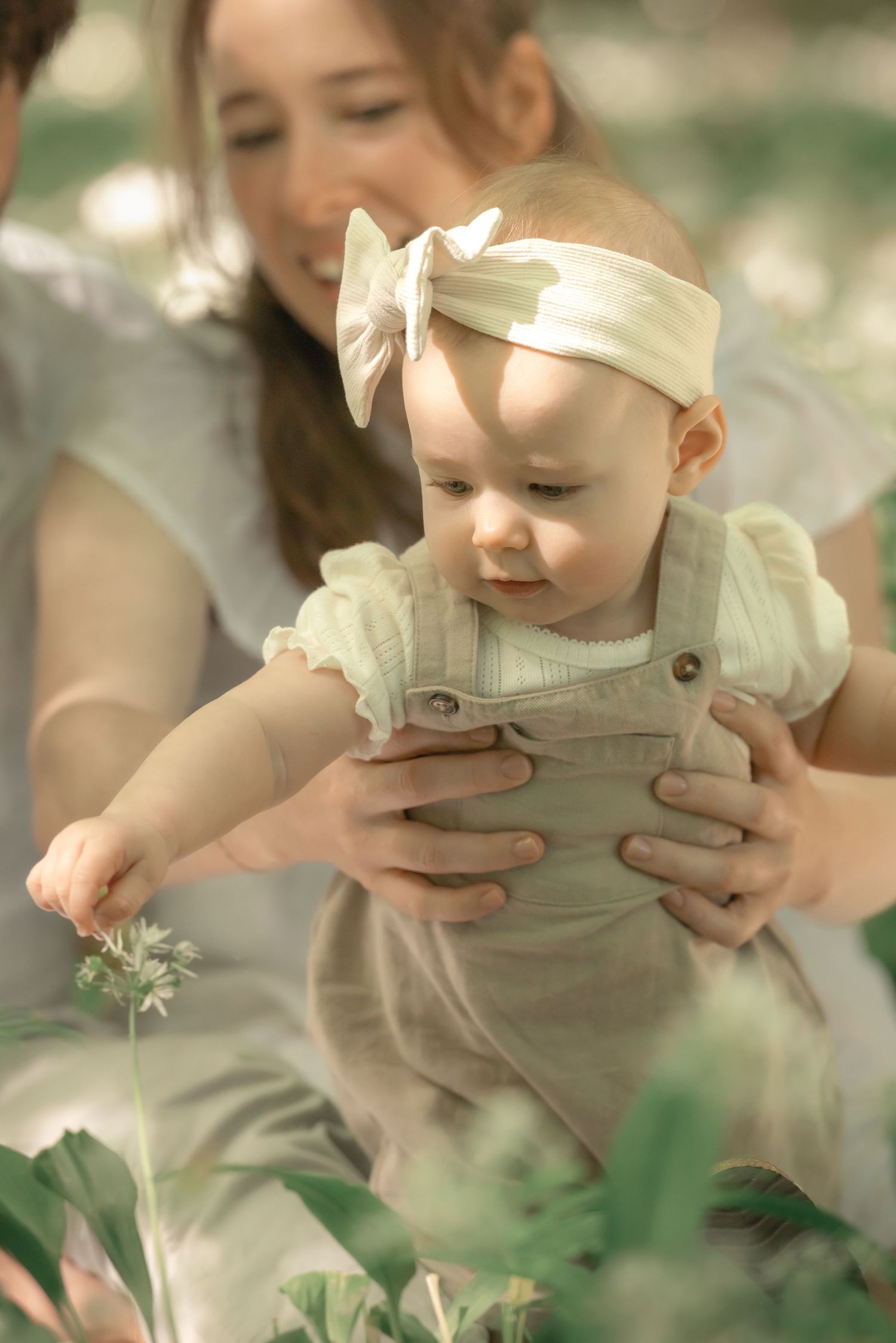Parents holding baby girl in linen overalls as she reaches toward white wildflowers in forest.
