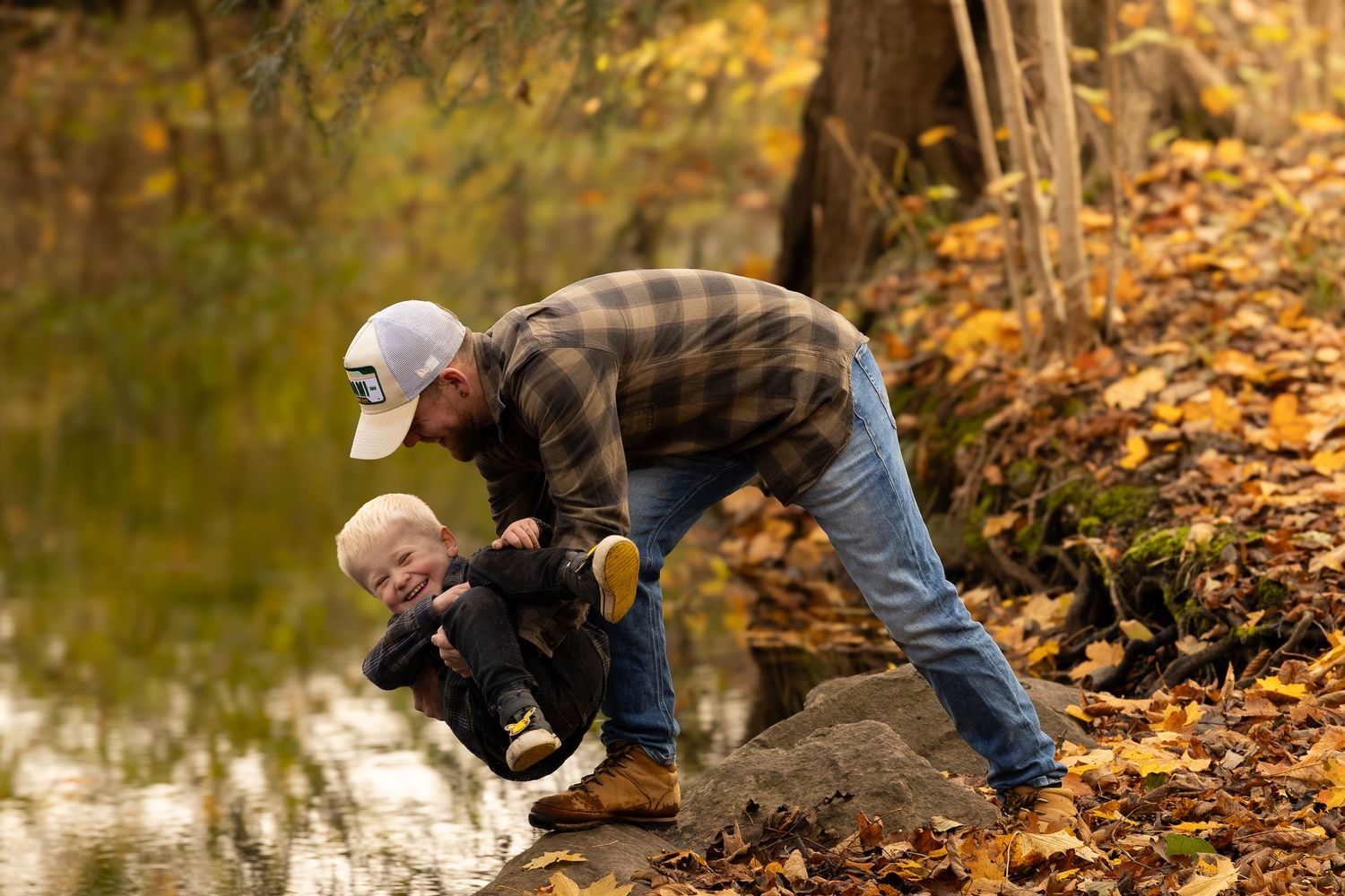 Father playfully bends down to interact with his toddler by a pond in an autumn woodland setting.