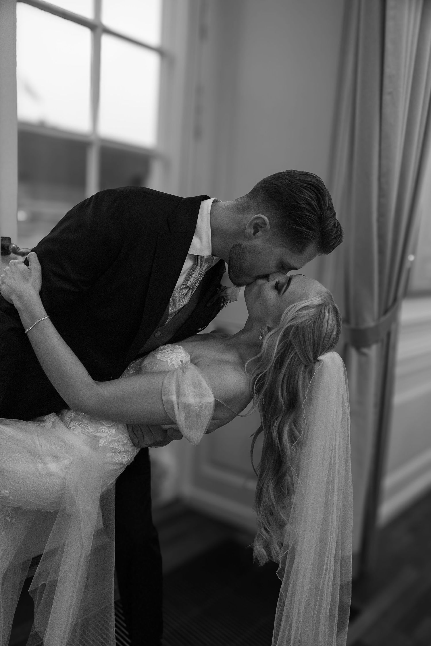 Romantic black and white close-up of bride and groom sharing a dip kiss by window.