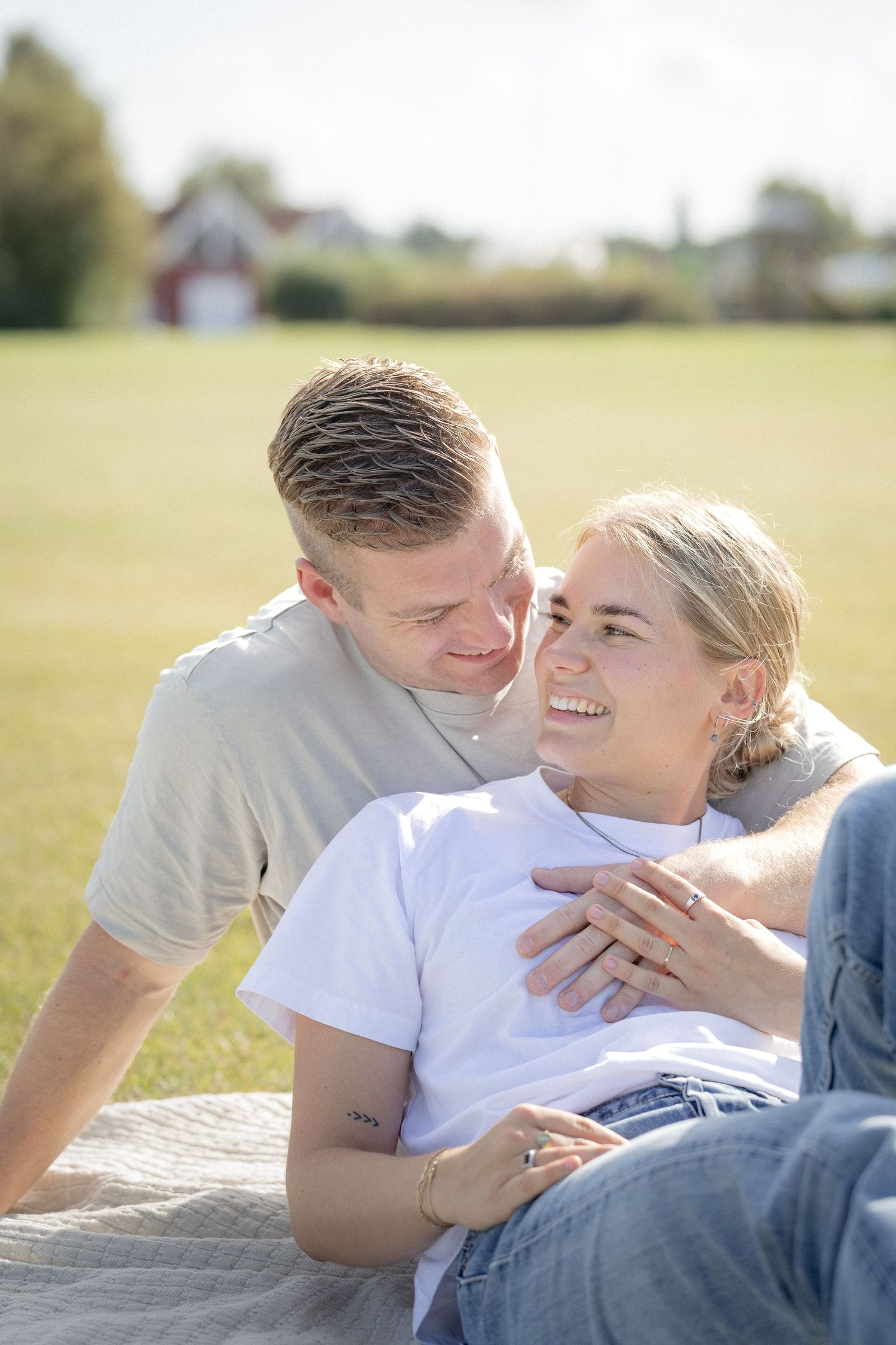 A happy young couple relaxes on a blanket in a sunny green field, smiling and holding each other closely.