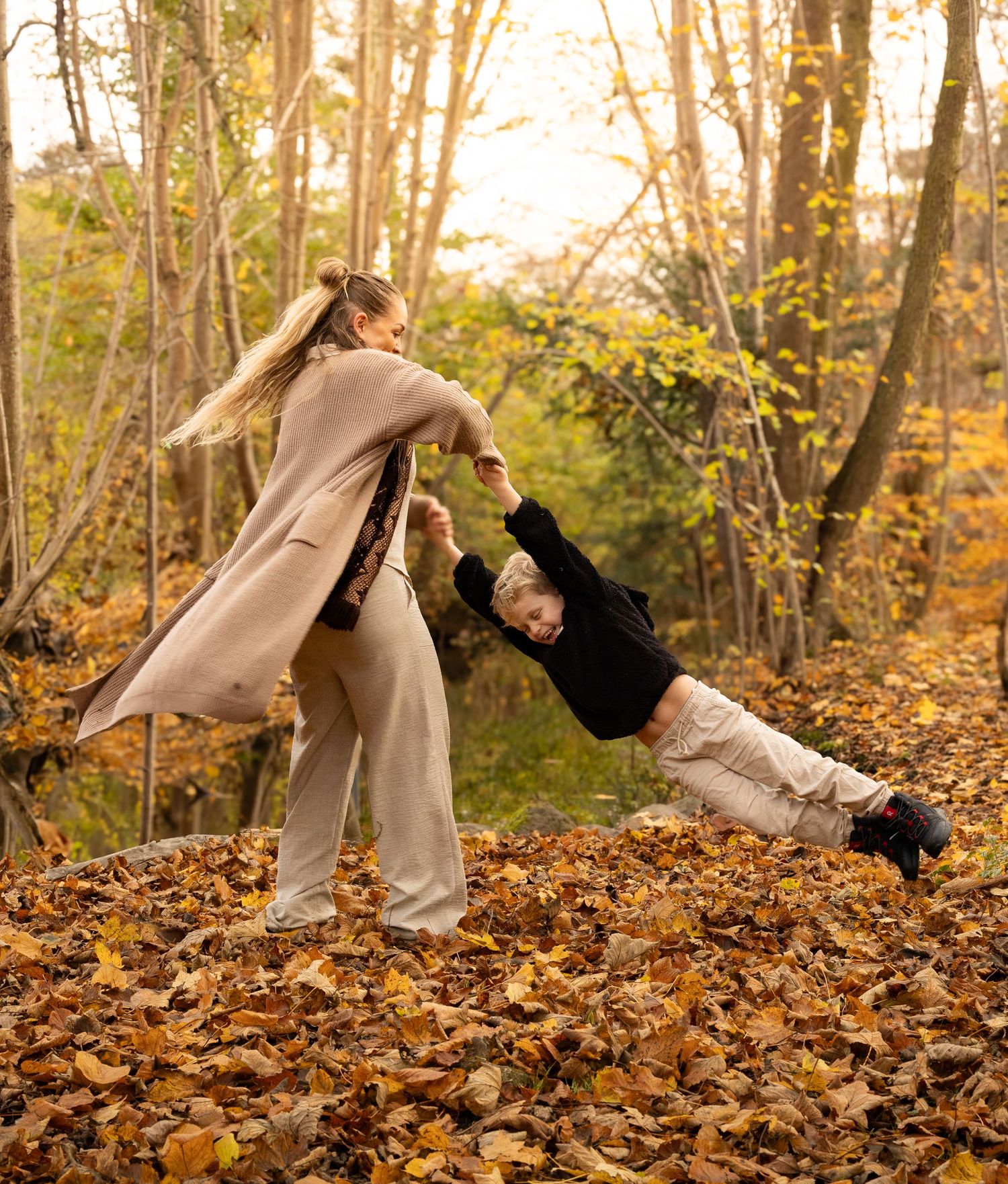 Mother swinging her child through colorful autumn leaves in a sunlit forest clearing.