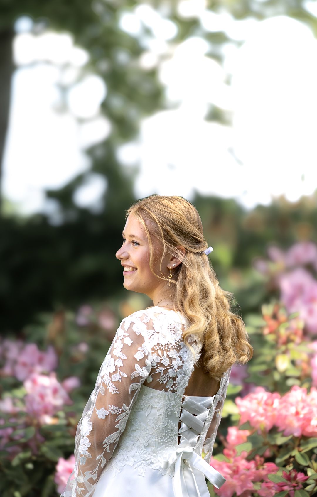 Blonde girl in white lace dress with open back smiling among pink flowering bushes.