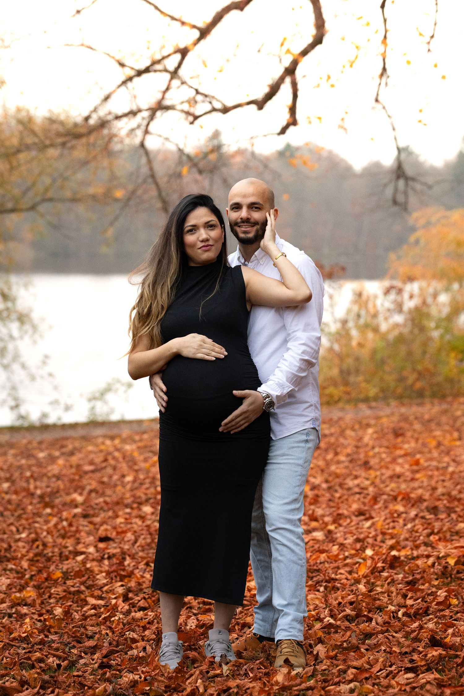 Expecting couple smiling together in autumn park with colorful fall foliage and lake in background.