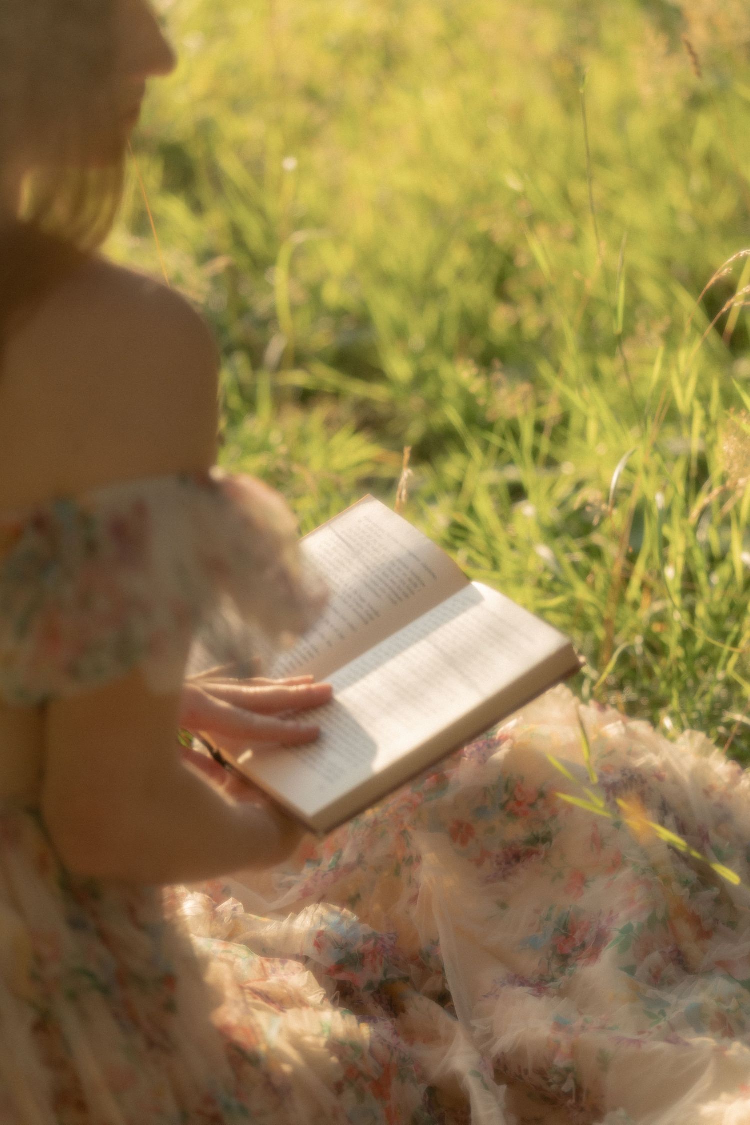 Woman in floral dress reading a book outdoors in a sunny meadow with green grass in the background.