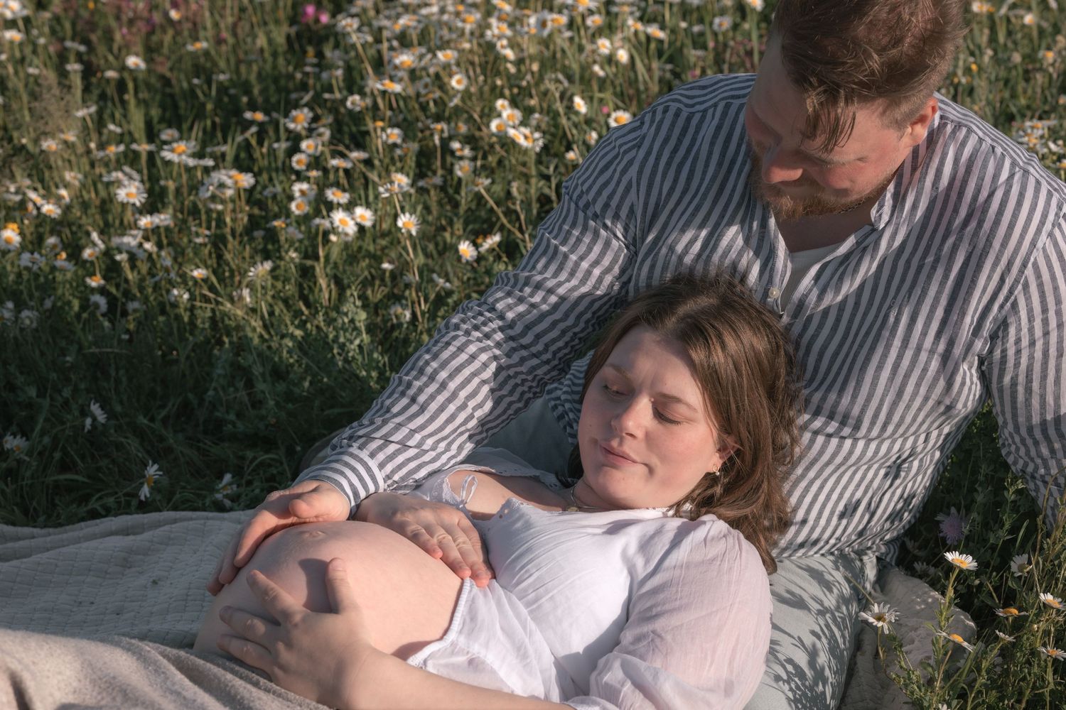 Couple relaxing in a daisy meadow, pregnant woman lying on partner's lap in warm sunlight.