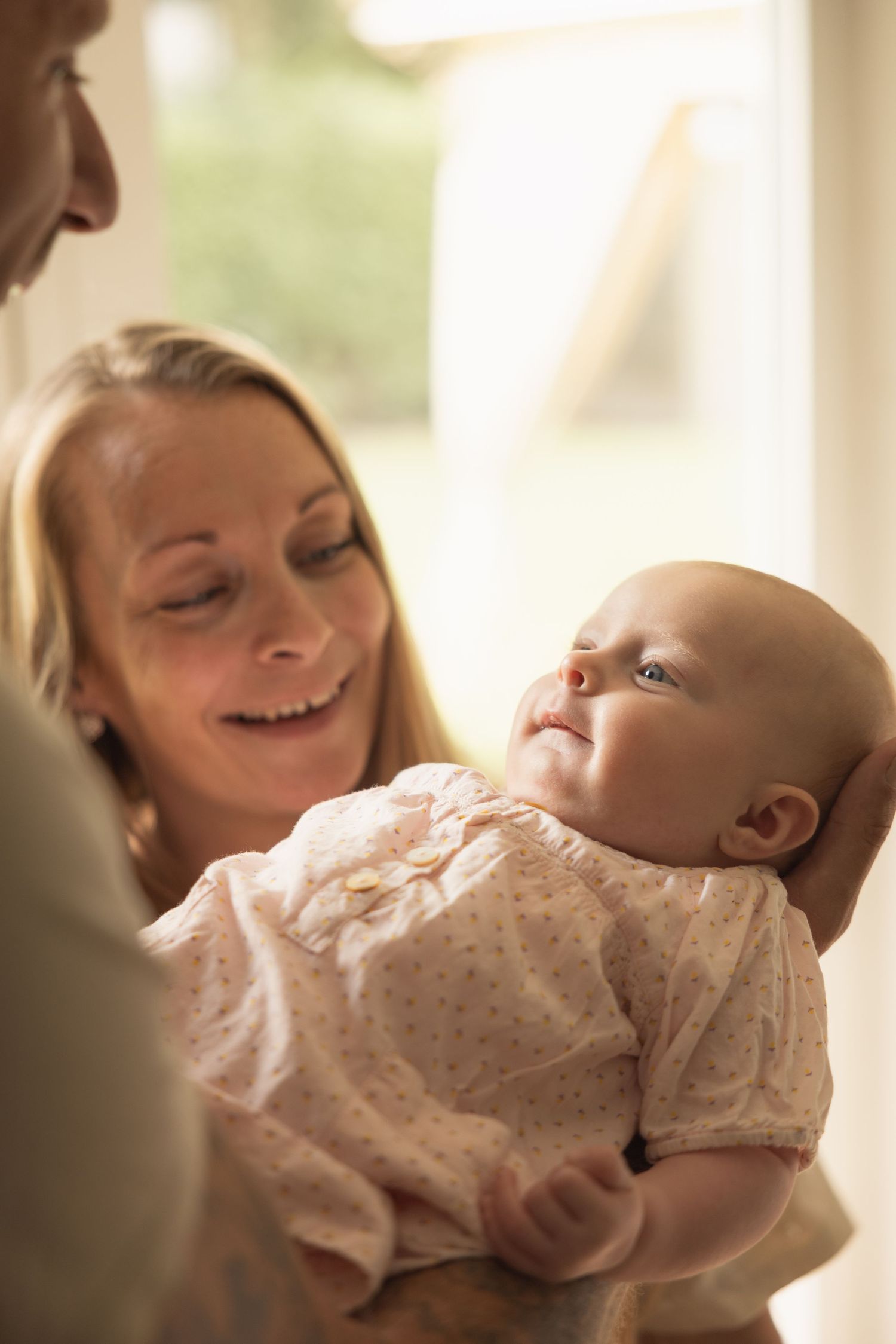 Smiling blonde mother holding her newborn baby in a pink outfit near a bright window.